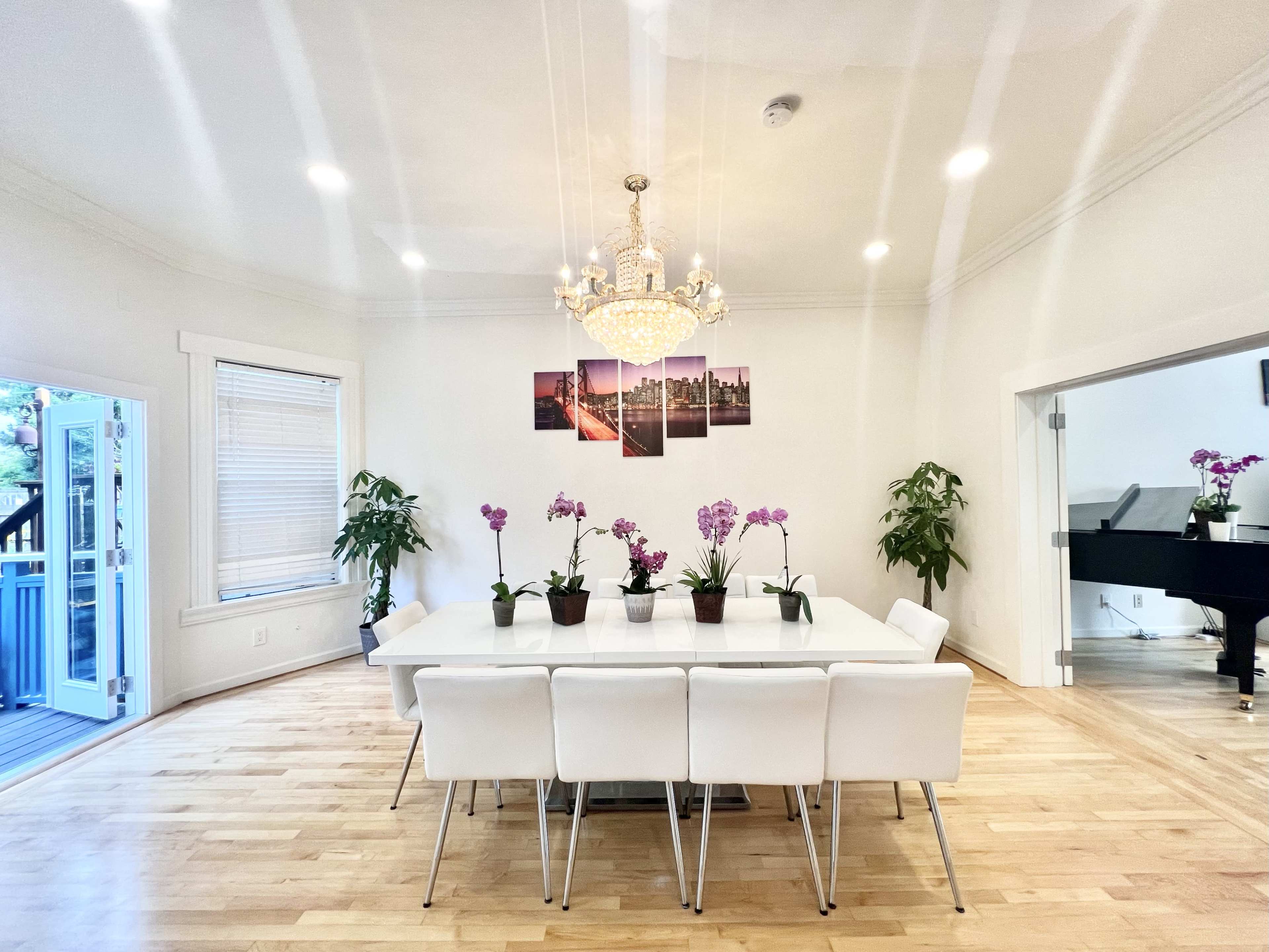 A modern dining room features a long white table surrounded by chairs, with plants and purple orchids on the table, and a chandelier overhead.