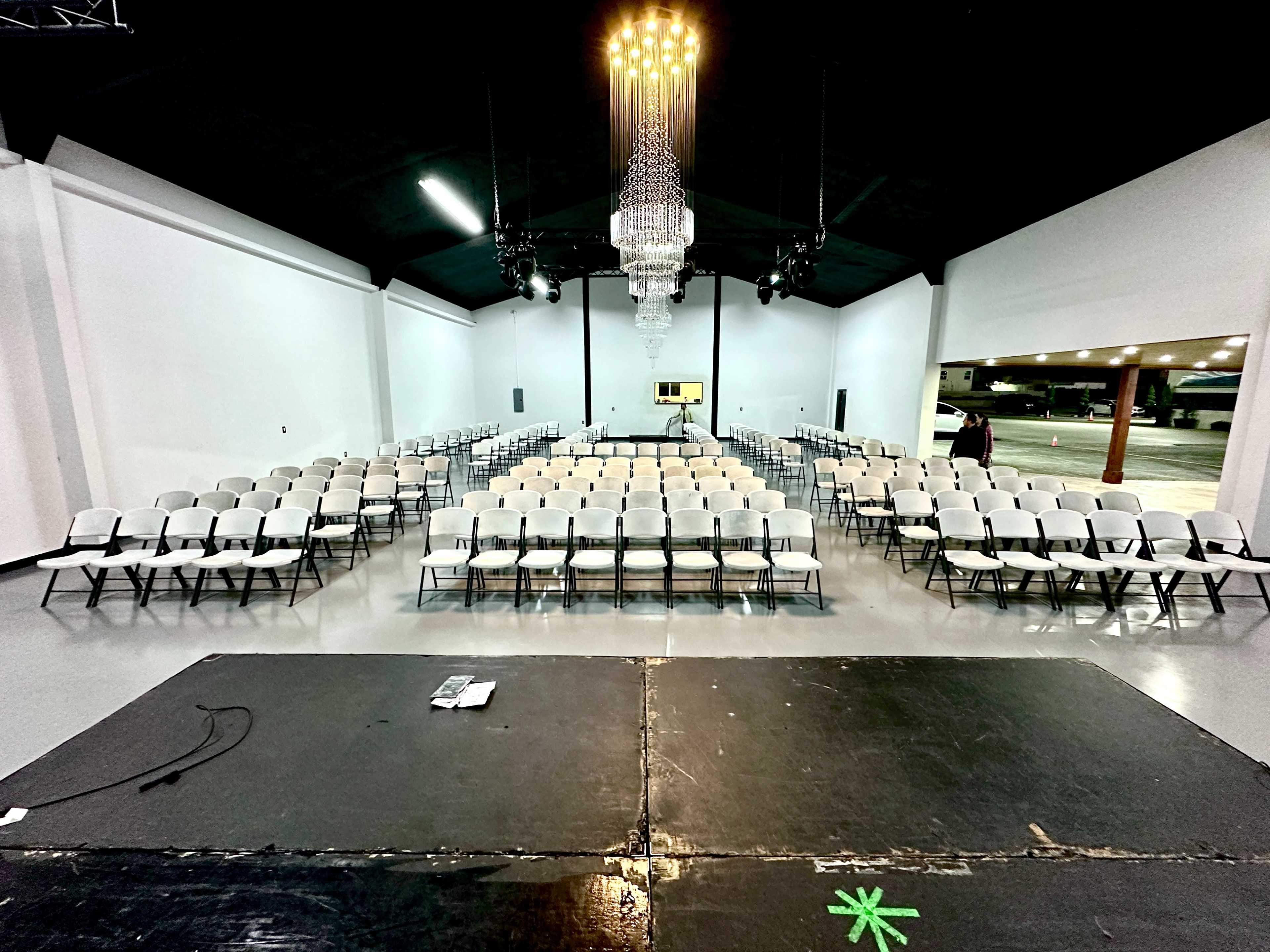 A spacious indoor venue set up with rows of white folding chairs facing a stage, illuminated by a chandelier overhead.
