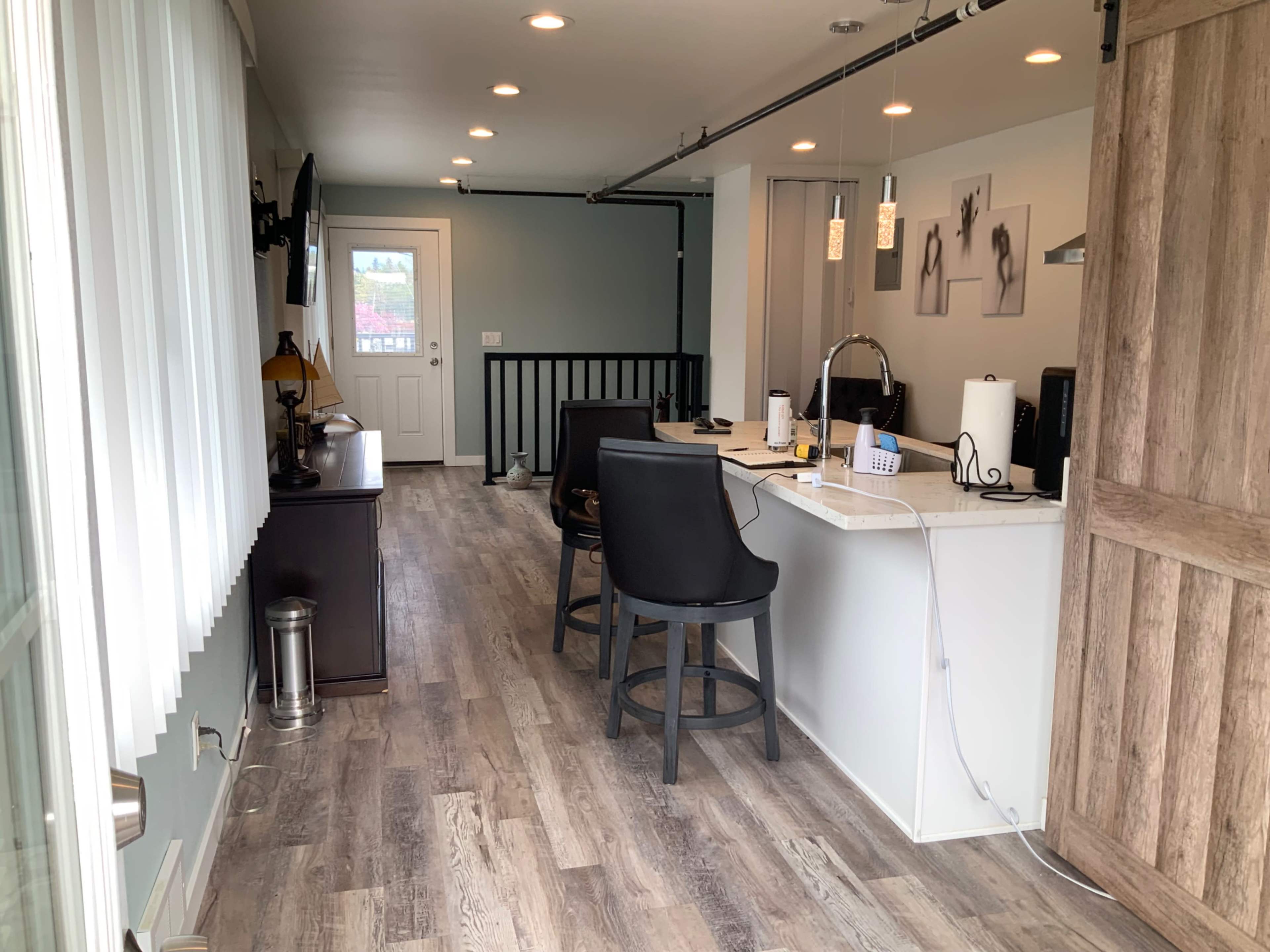 A spacious, modern interior with a kitchen bar area, black stools, a door leading outside, and large windows allowing natural light.