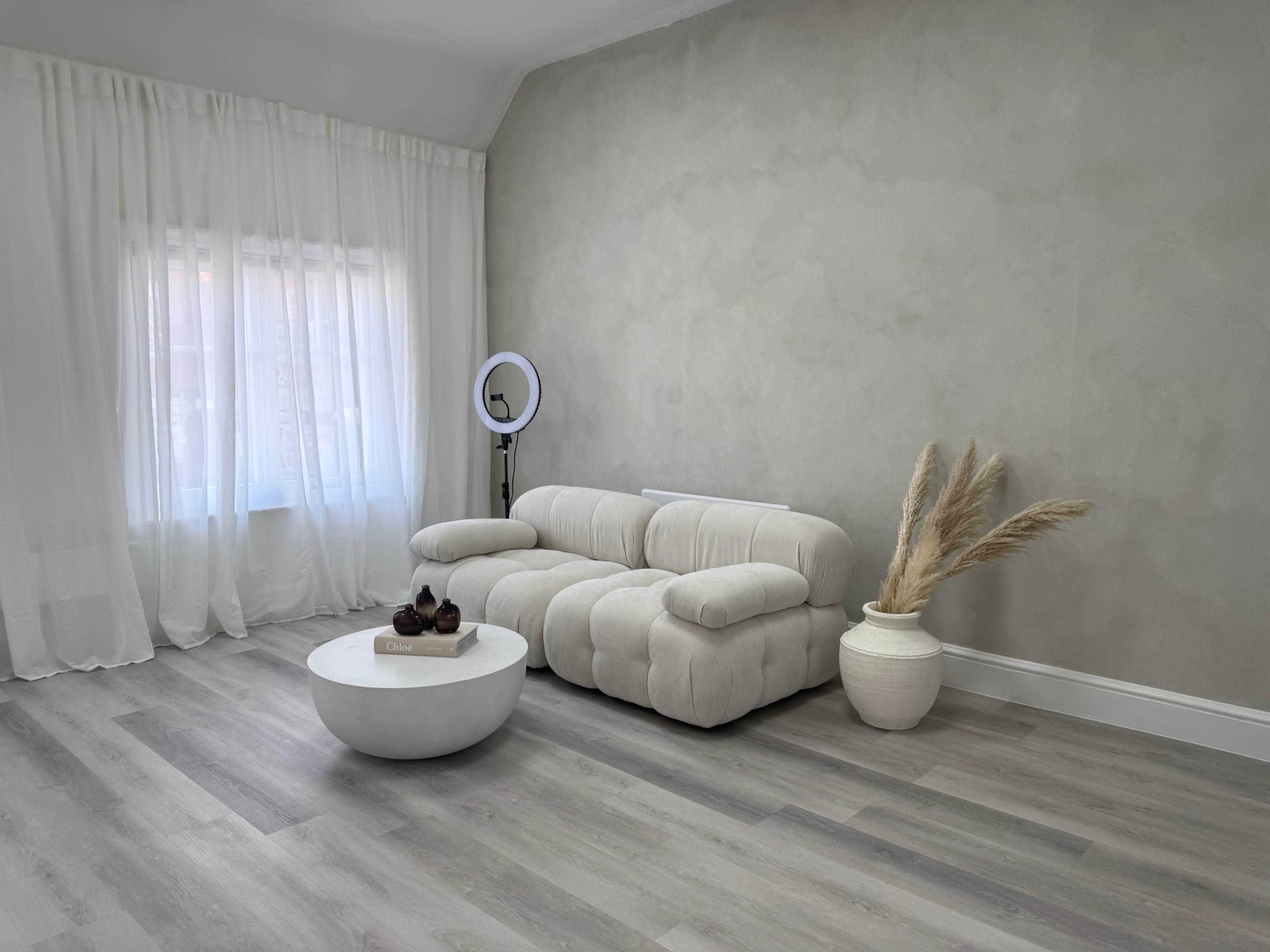 The image shows a modern living room with a curved beige sofa, a circular white coffee table, and decorative elements like a vase and dried plants against a light-colored wall.