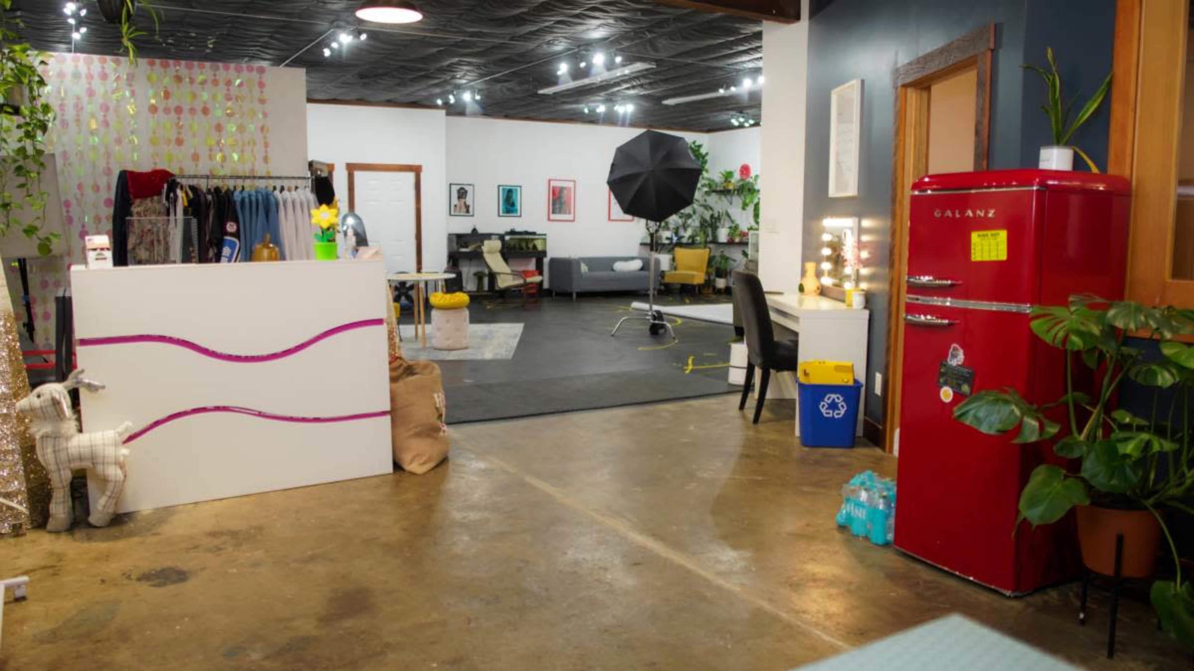 The image shows a bright studio space featuring a reception area with a red refrigerator, a clothing display, and a photo shoot setup in the background.