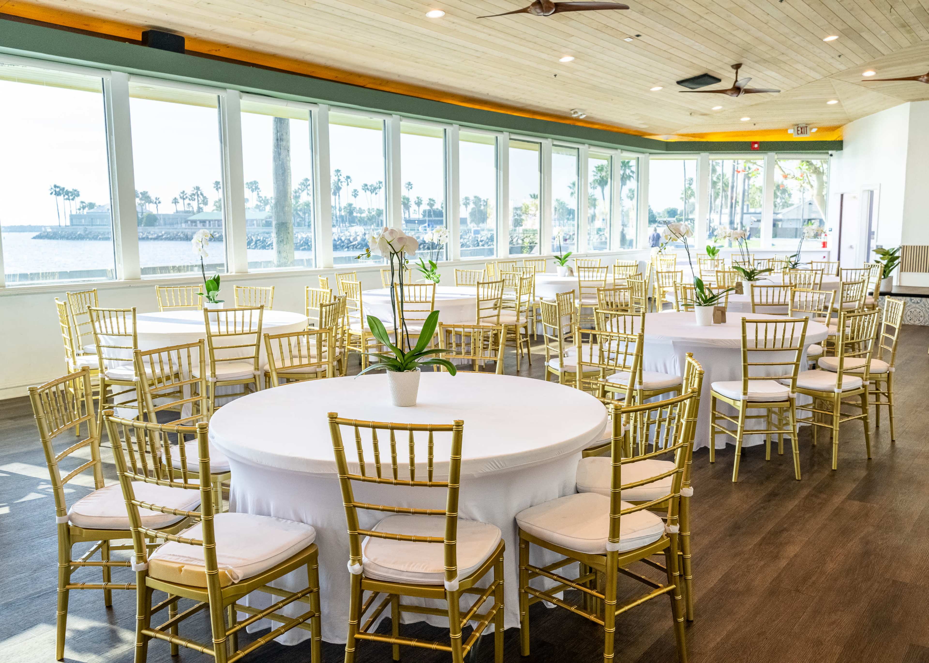 The image shows a banquet hall with round tables dressed in white tablecloths and gold chairs, featuring large windows that overlook a waterfront view.