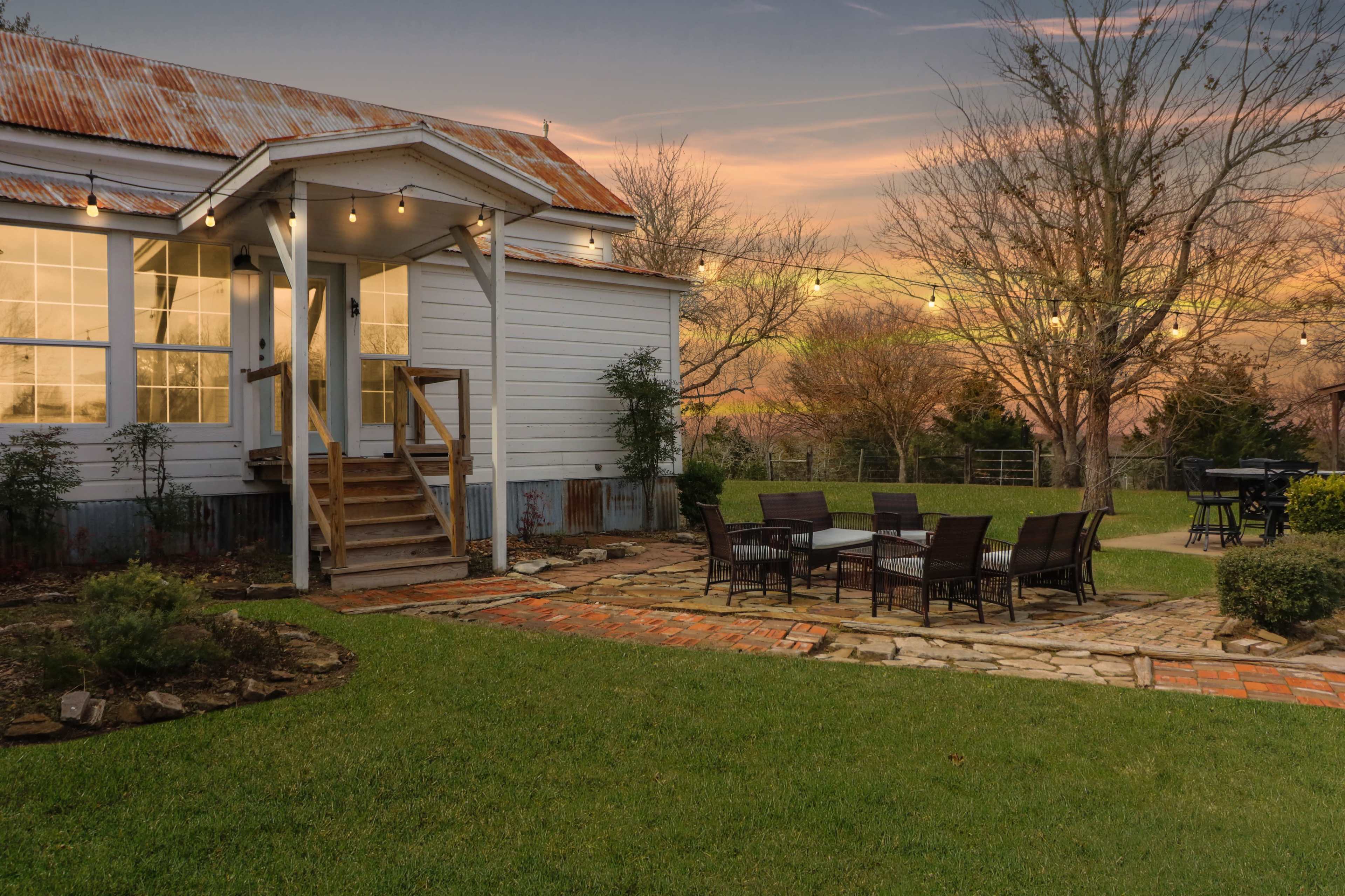 A house with a covered porch, a brick pathway, and a patio area with a stone seating arrangement in a grassy yard at dusk.