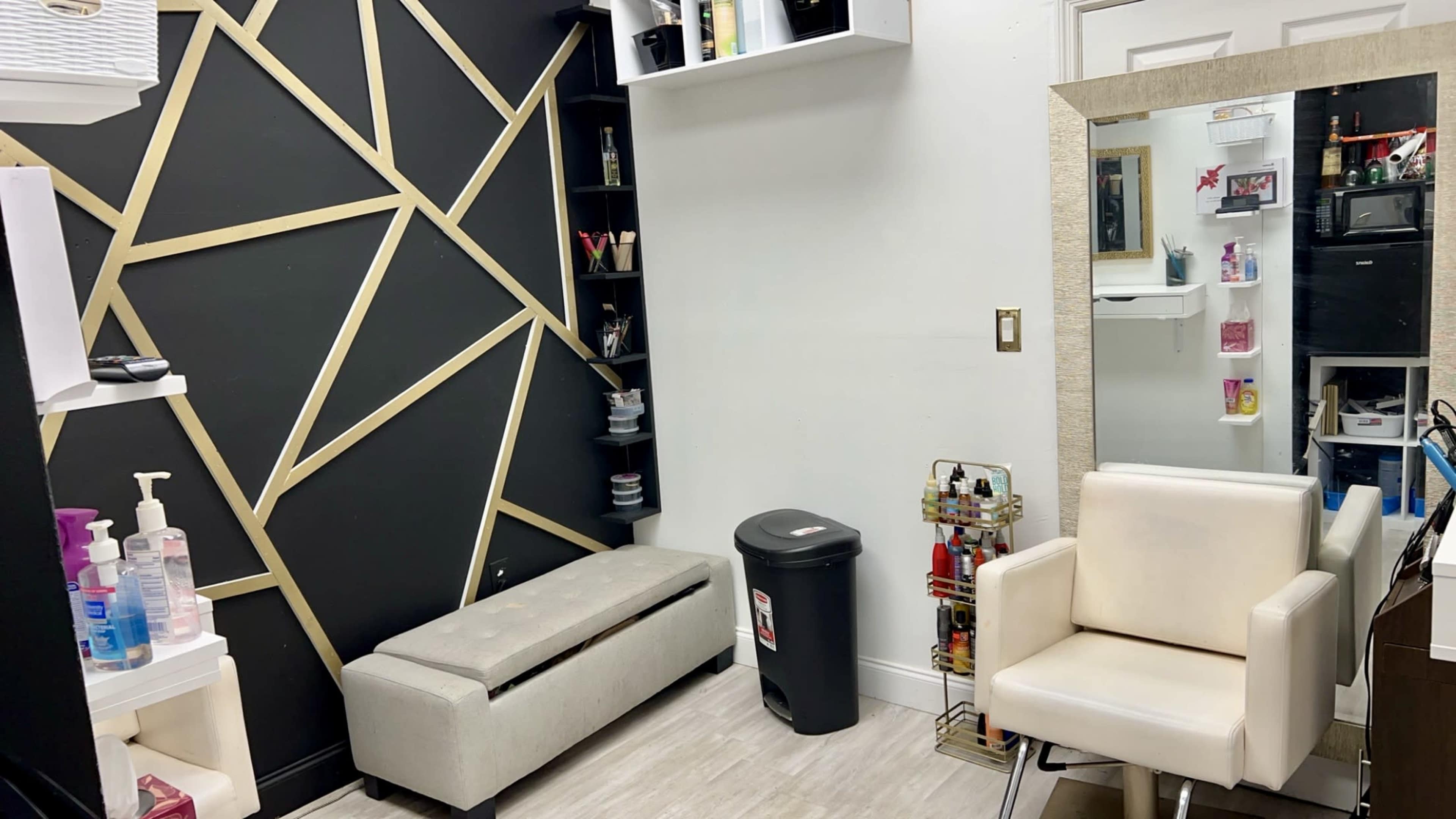 A minimalist salon space featuring a black and gold decorative wall, a white mirror, a gray bench, a white chair, and shelving with cosmetic products.