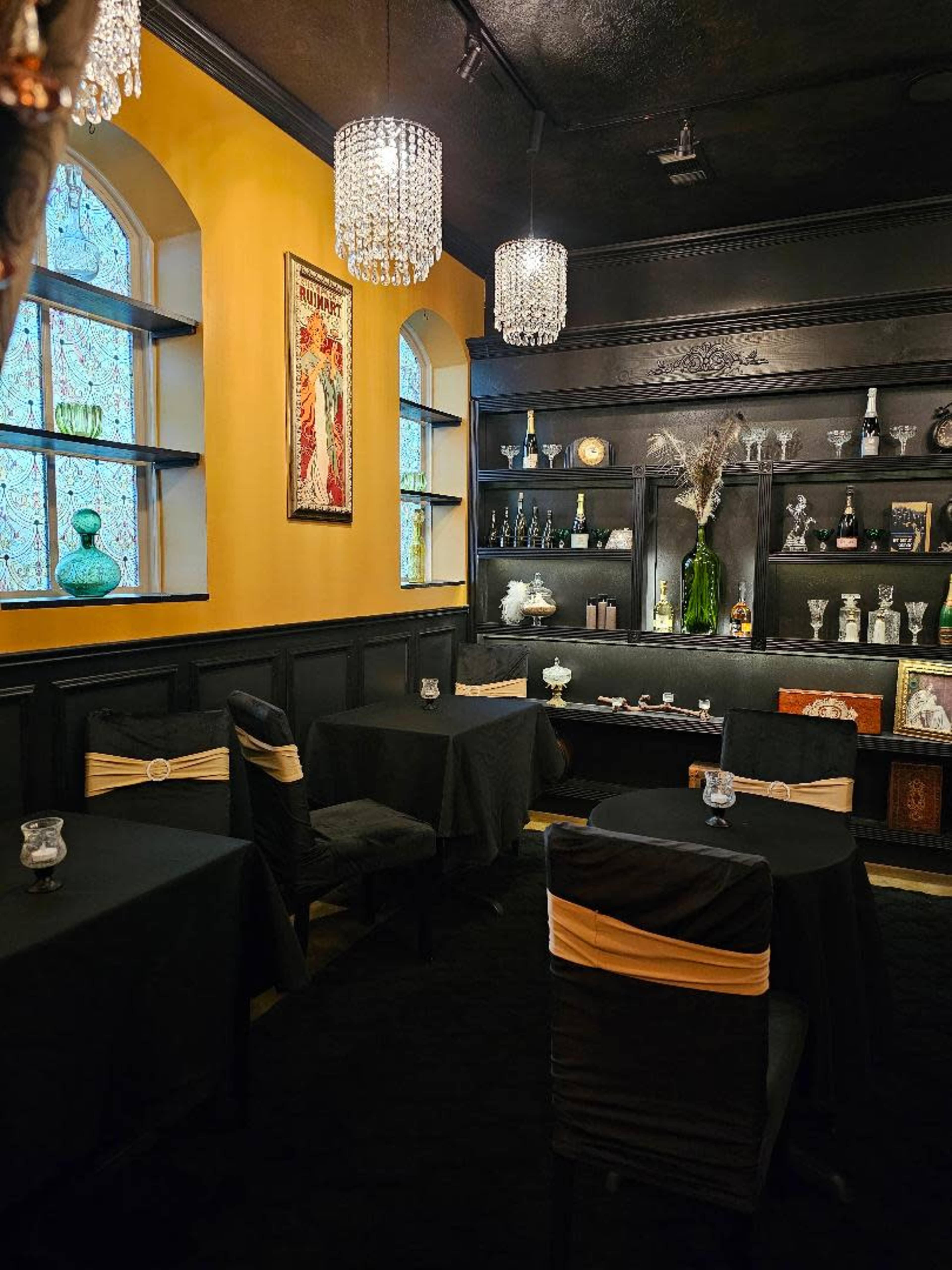The image shows a dimly lit restaurant interior with black tables and chairs, adorned with gold sashes, and shelves displaying decorative bottles and glassware against a yellow wall.