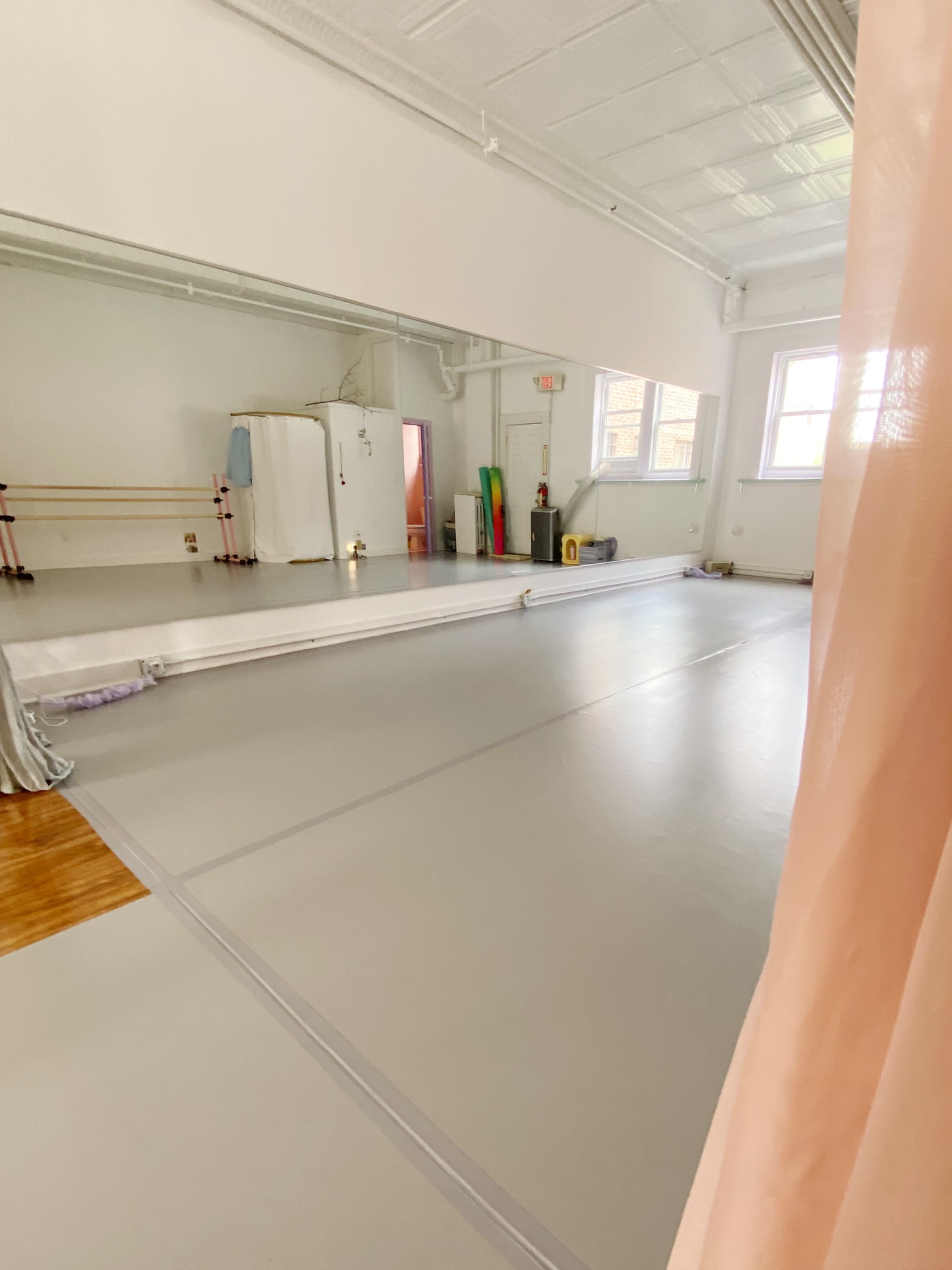 The image shows a spacious dance studio with a mirrored wall, wooden ballet barres, and a gray floor.