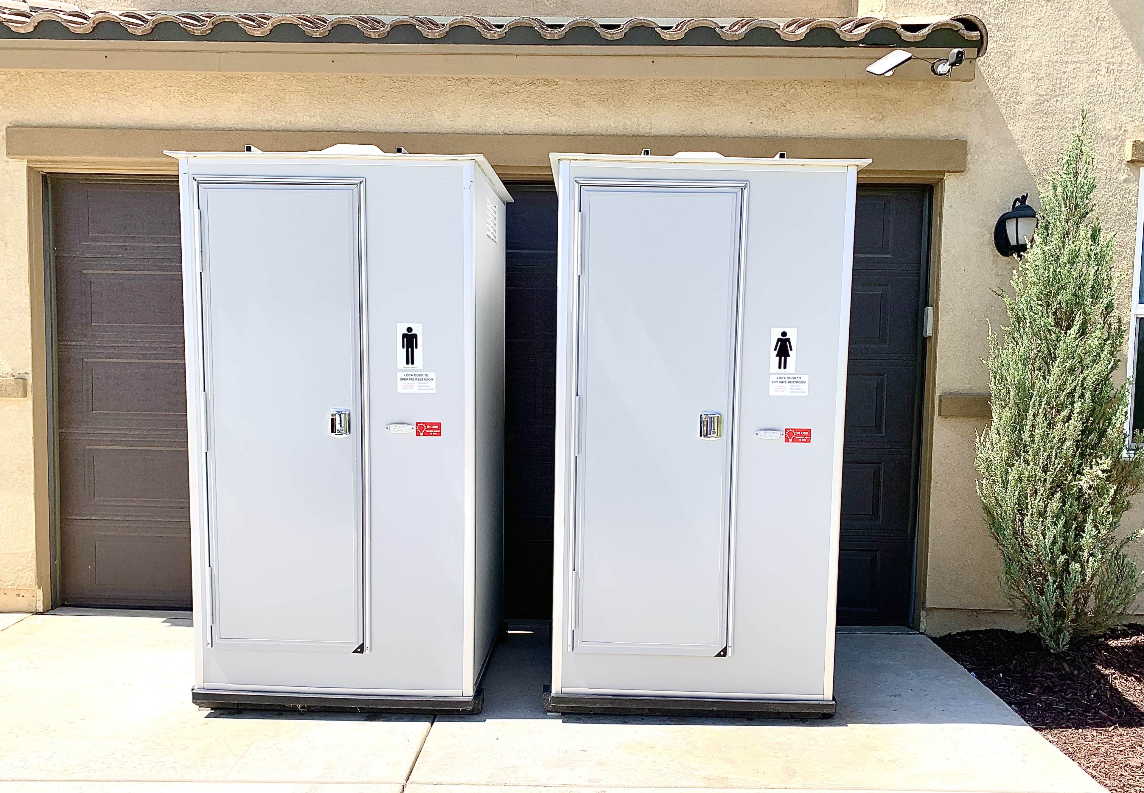 Two portable restroom units are positioned next to each other in front of a building with dark garage doors.