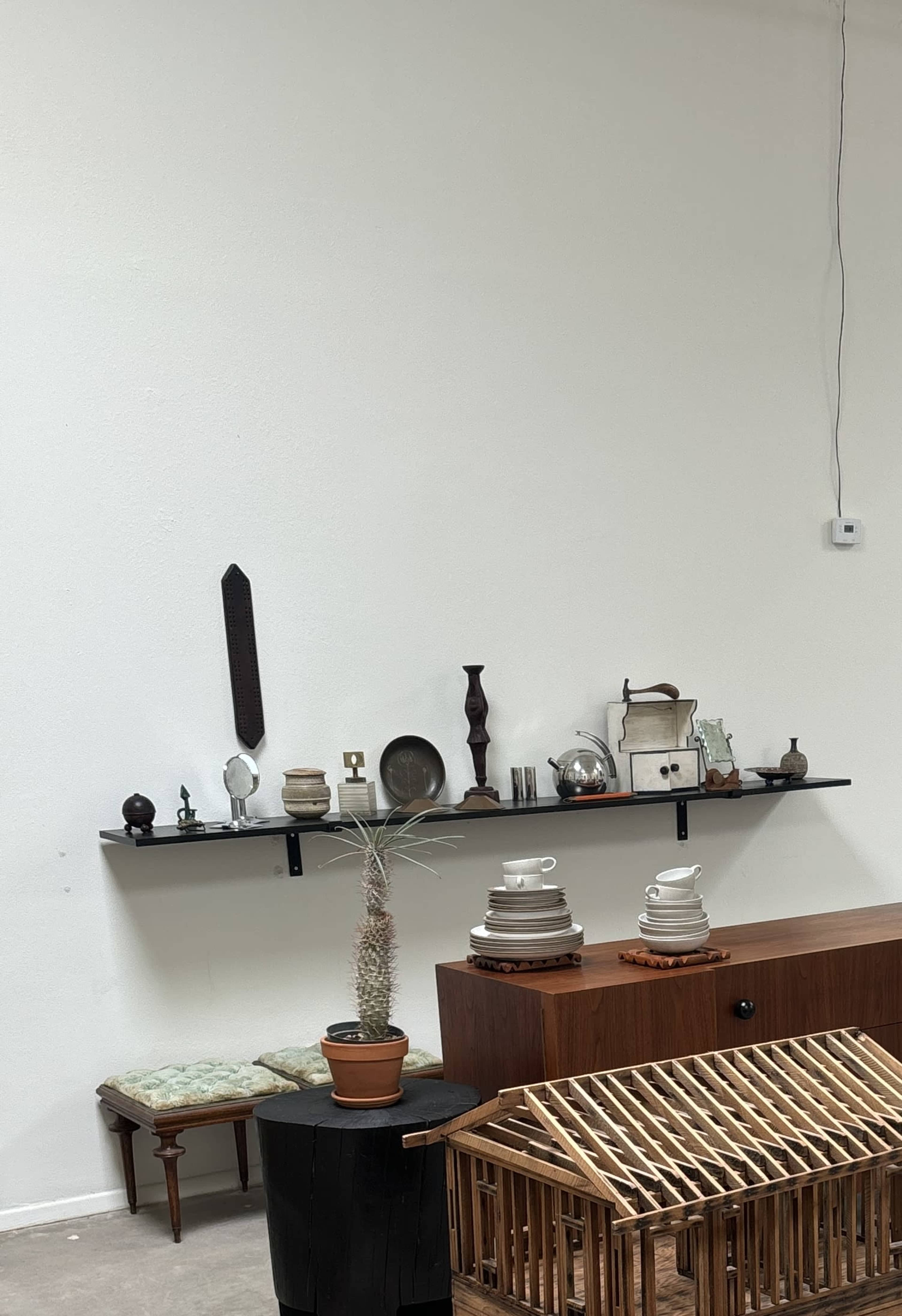 A wall with a black shelf displaying various ceramic and metal objects, a wooden cabinet, a potted plant, and a wooden structure in the foreground.