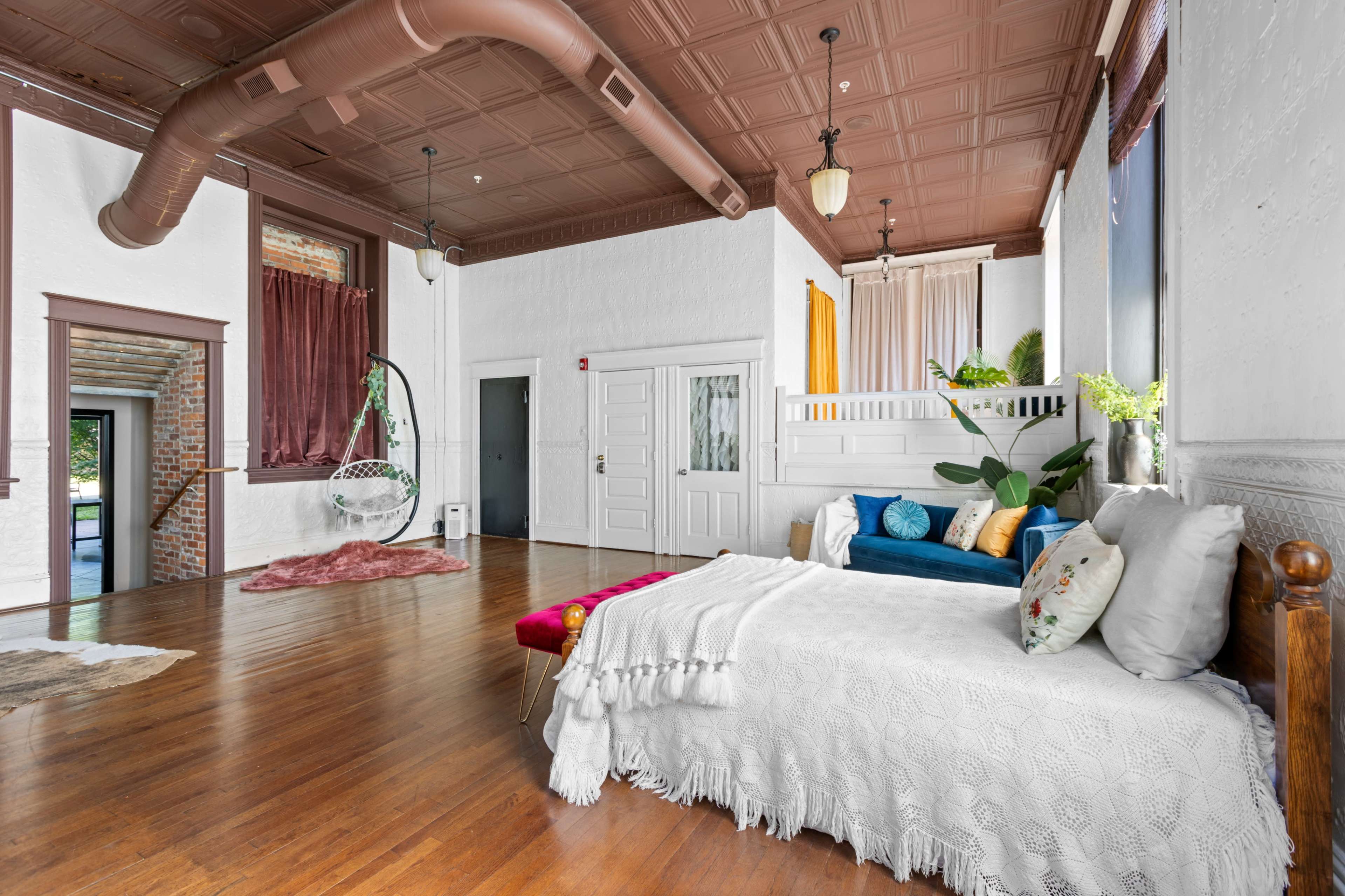 The image shows a spacious interior with hardwood floors, a daybed, and a seating area, highlighted by a ceiling with decorative tiles and large windows.