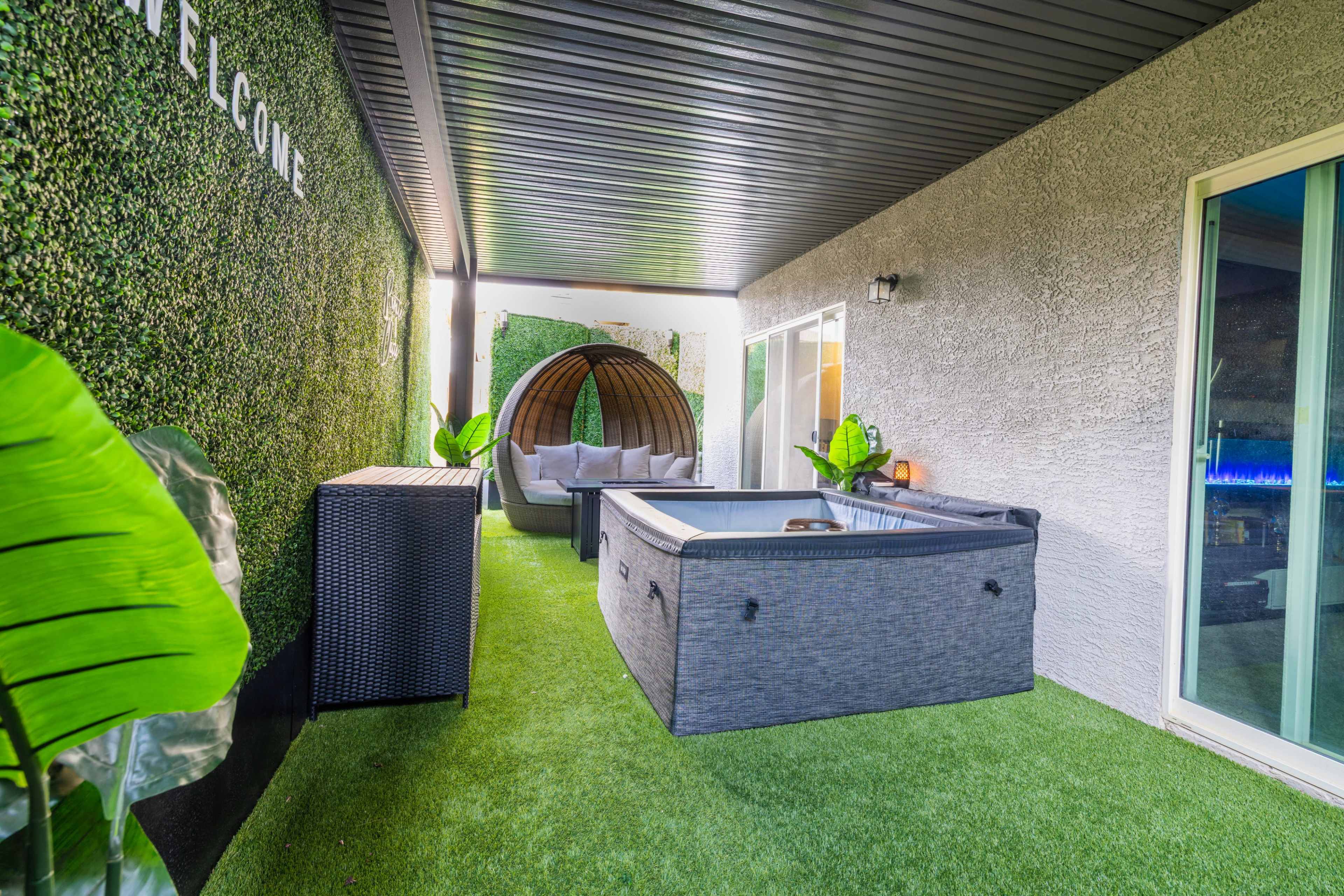 The image shows a modern outdoor patio area featuring a hot tub, a lounge seating arrangement, and decorative plants set against a backdrop of artificial grass and a green wall.