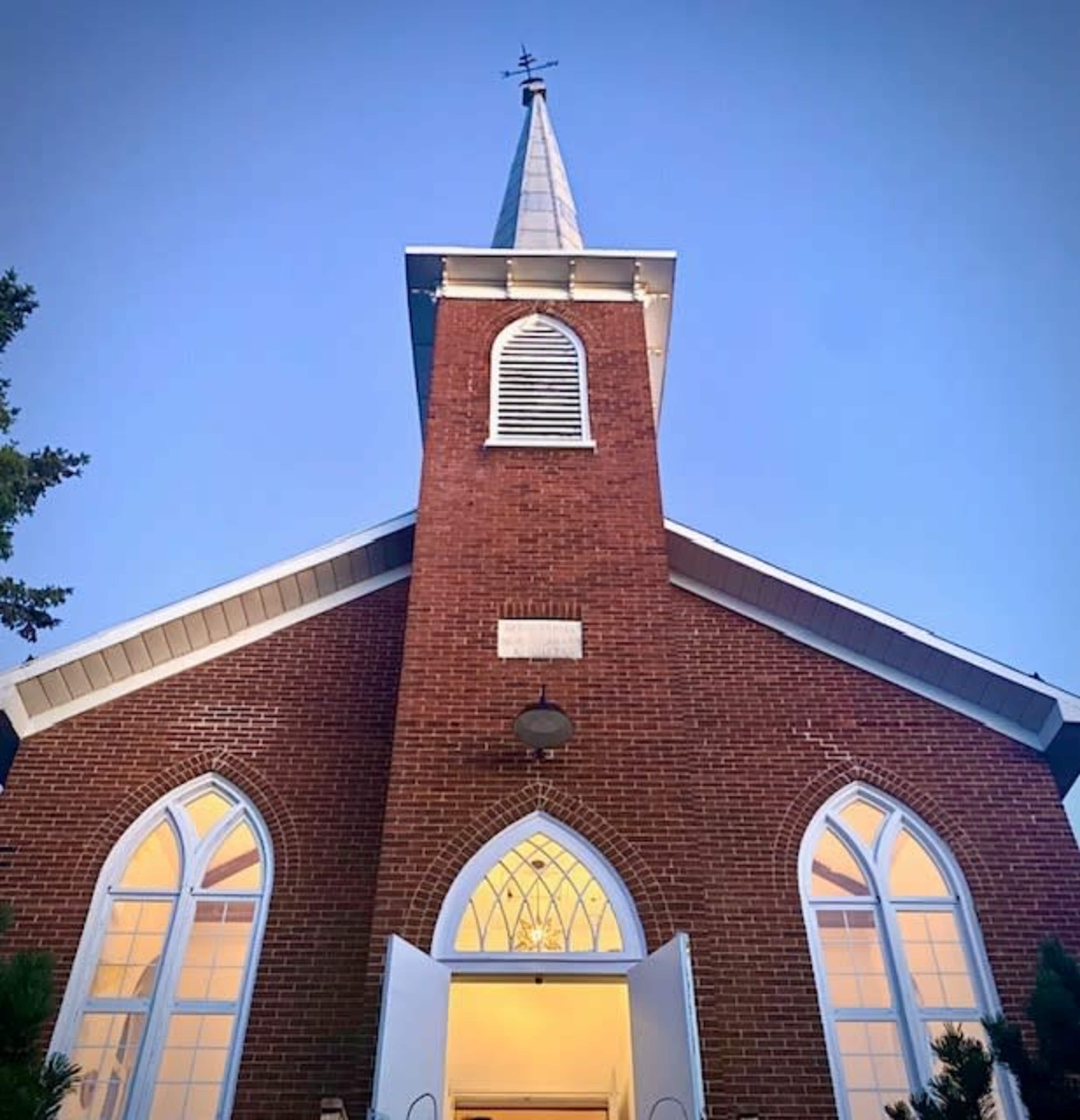 A brick church with a tall steeple and arched windows is illuminated at dusk.