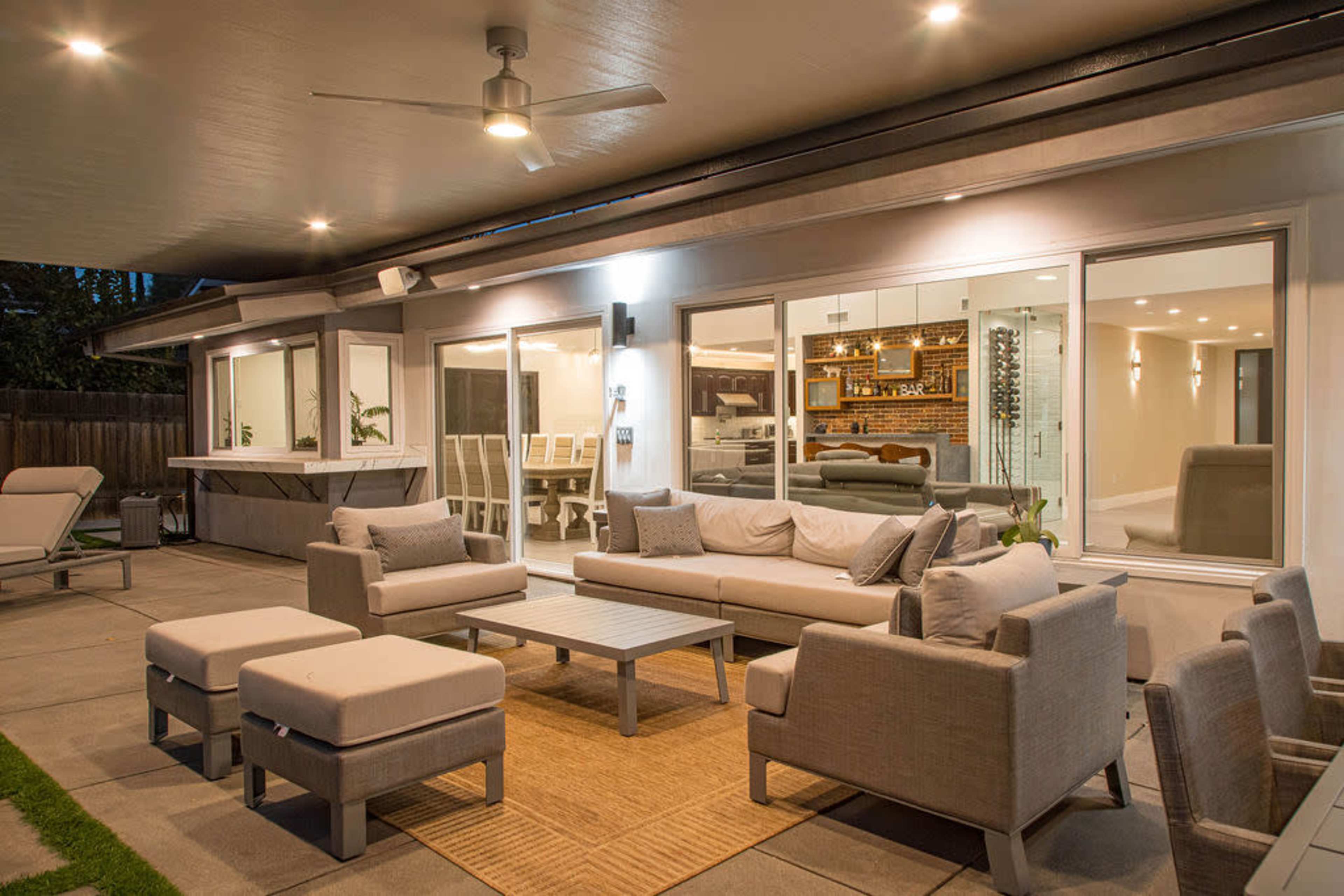 A modern outdoor seating area features comfortable gray furniture arranged around a coffee table, overlooking large windows that provide a view of an interior dining space.