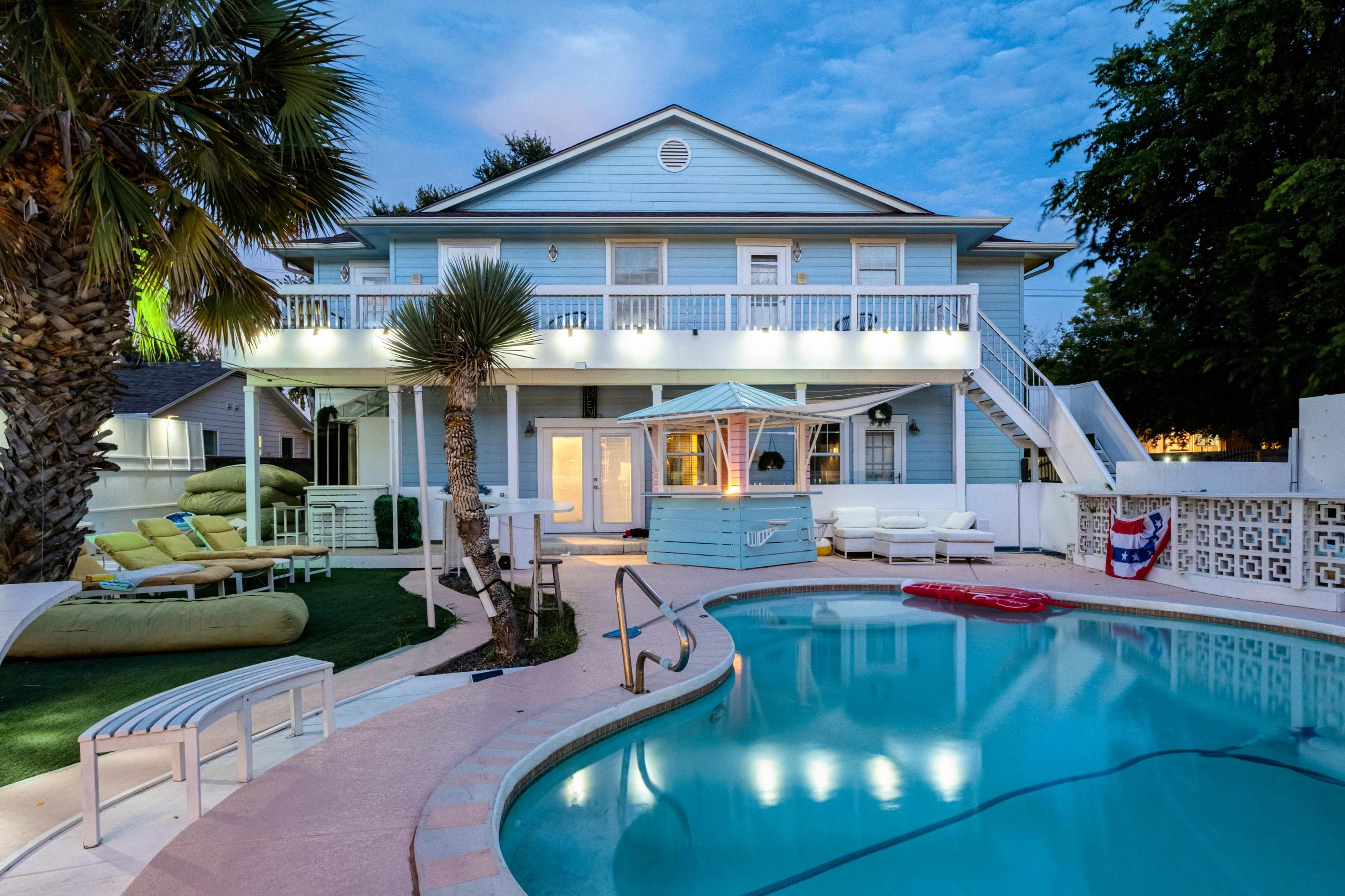 The image shows a two-story light blue house with a swimming pool and lounge area in the foreground, surrounded by palm trees and outdoor furniture.