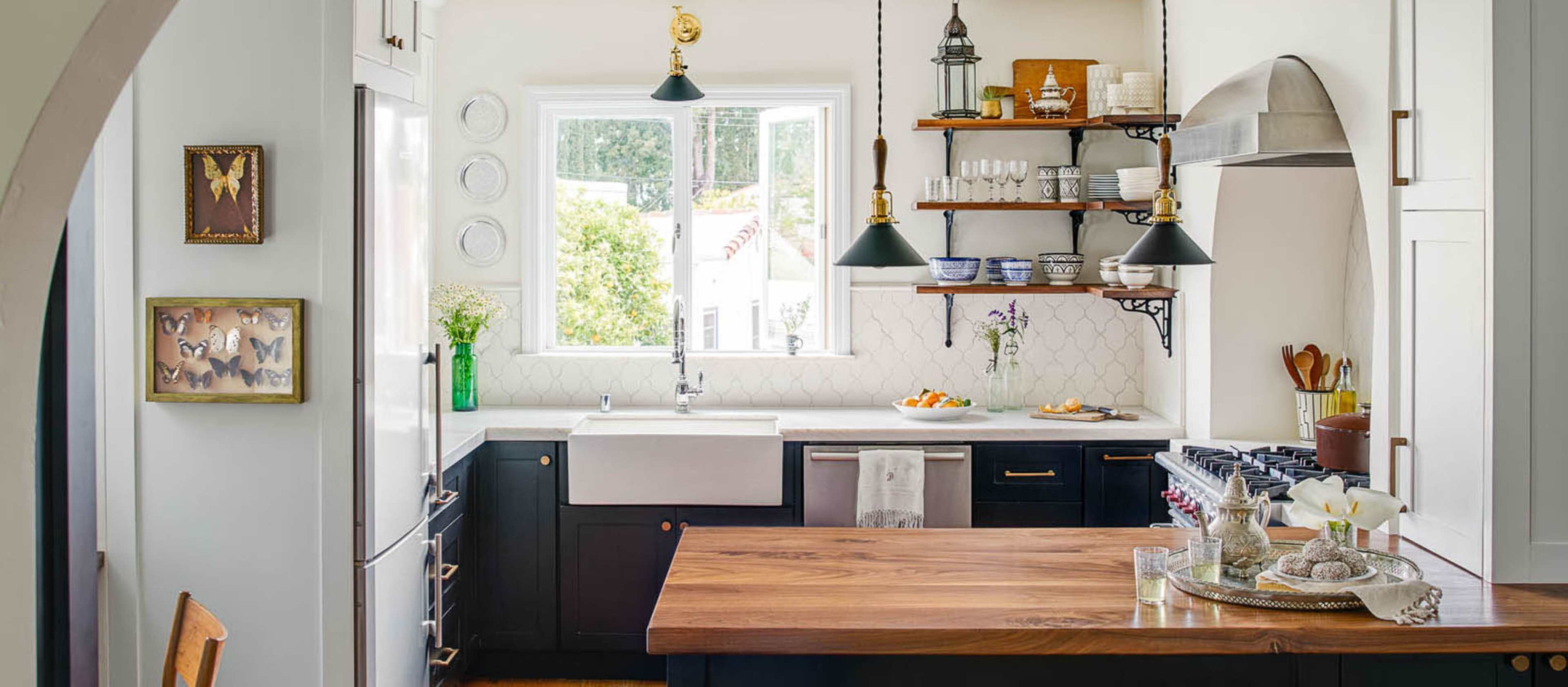 A modern kitchen features a large wooden island, dark cabinetry, open shelving with dishware, and a window overlooking an outdoor view.