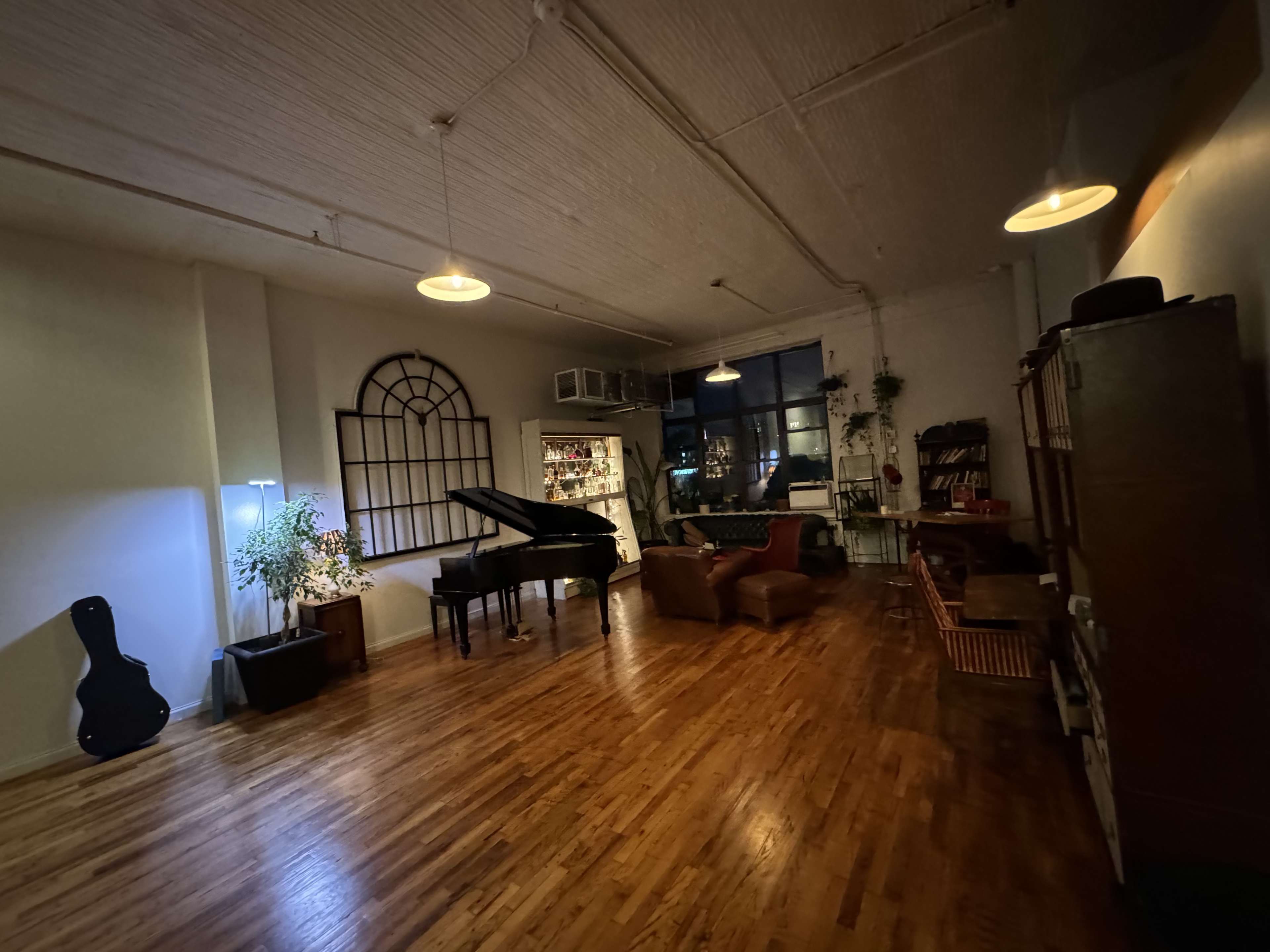 The image shows a spacious, dimly lit room featuring a grand piano, a guitar resting against a plant, and various pieces of furniture along the walls.
