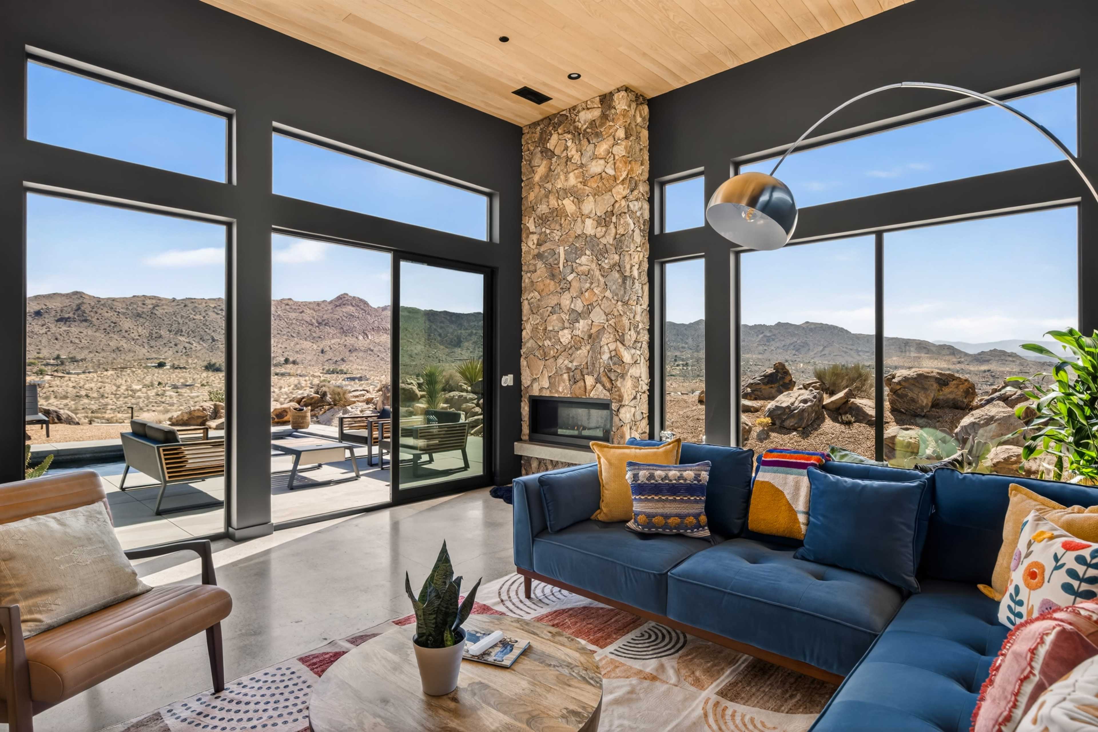 The image shows a modern living room with large windows overlooking a desert landscape, featuring a blue sofa and a stone fireplace.