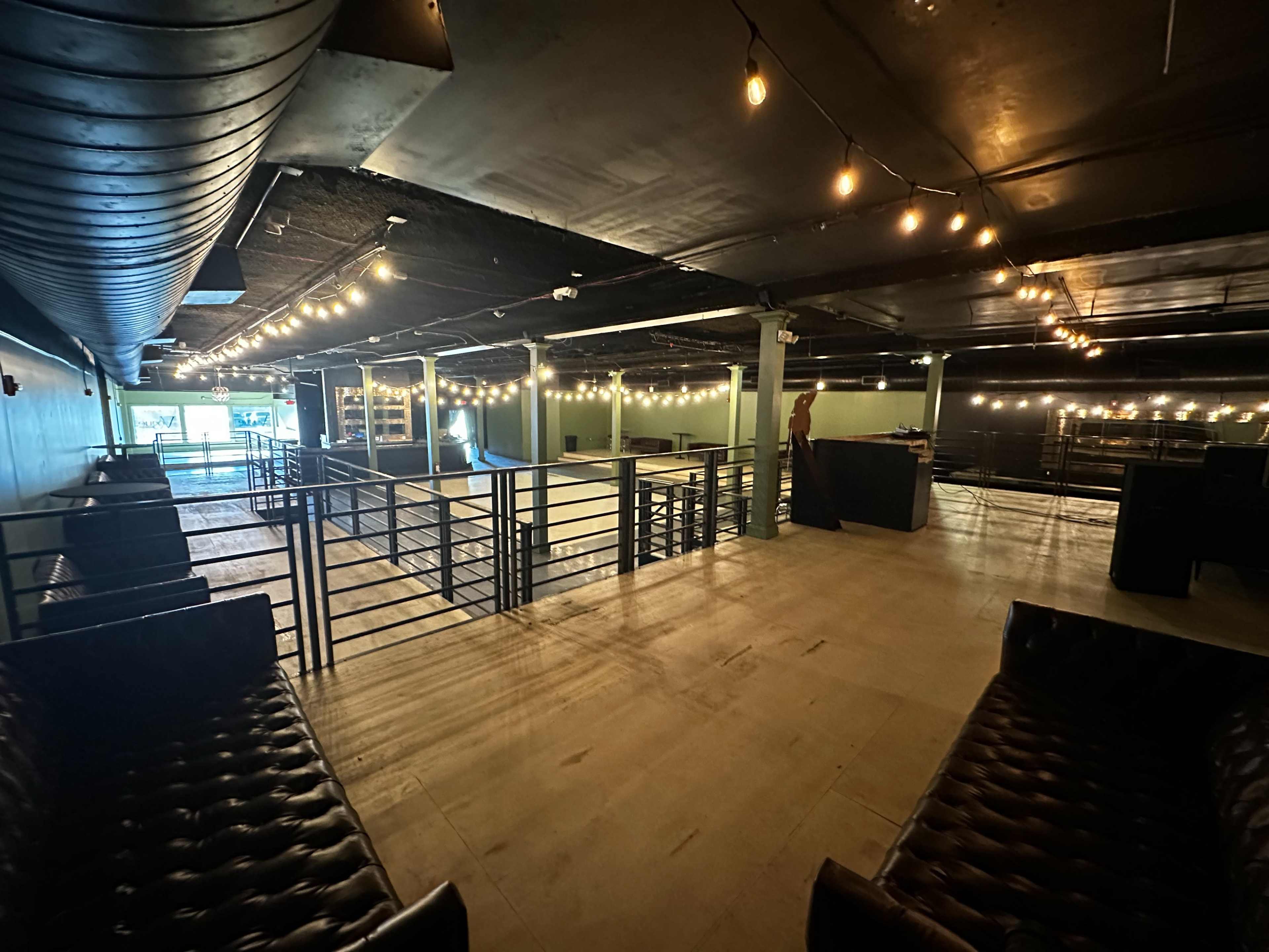 The image shows an empty, well-lit indoor venue with string lights and seating areas arranged around a central space.