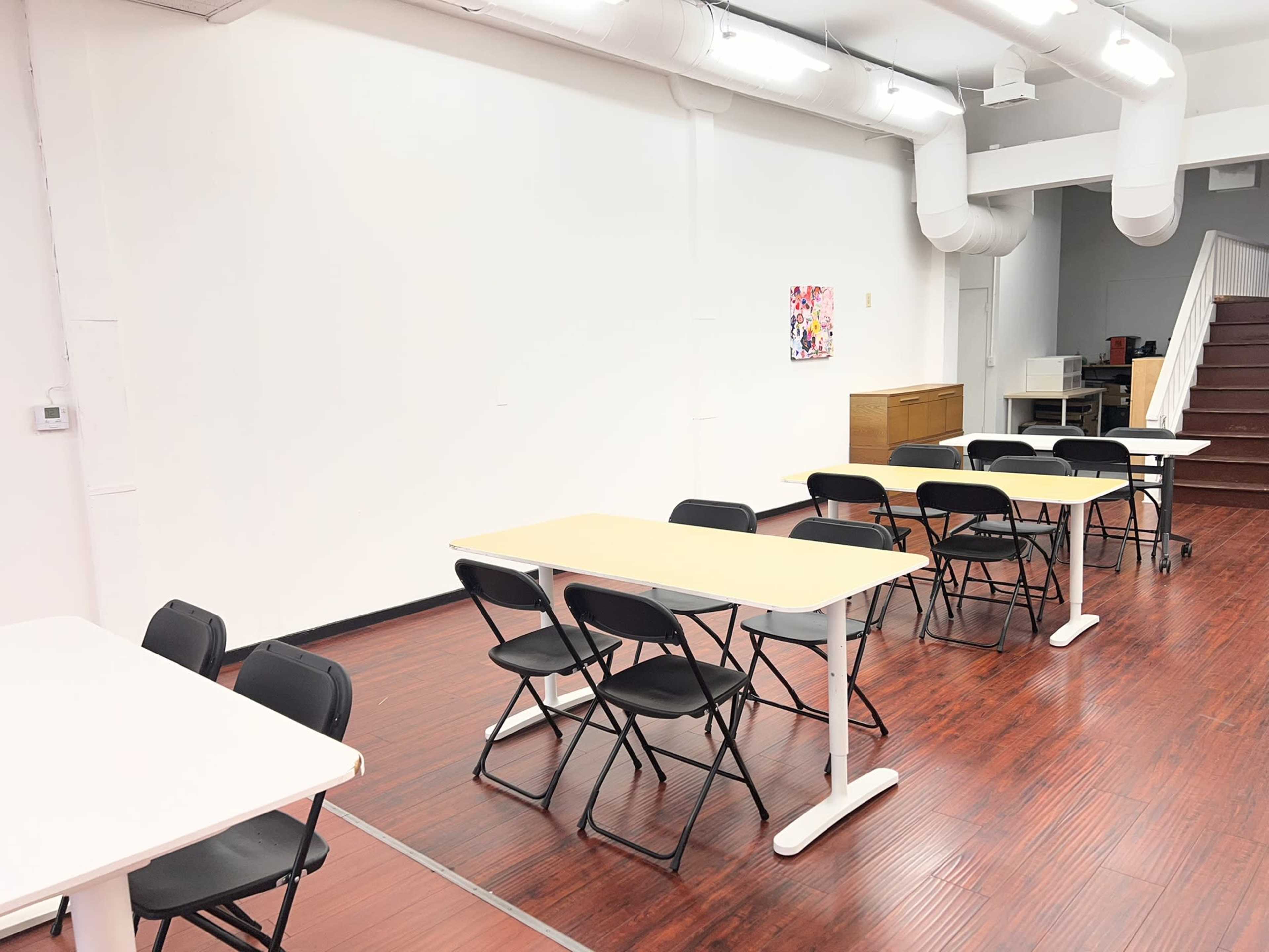 The image depicts a spacious, well-lit classroom with several folding tables and chairs arranged neatly, and a staircase visible in the background.