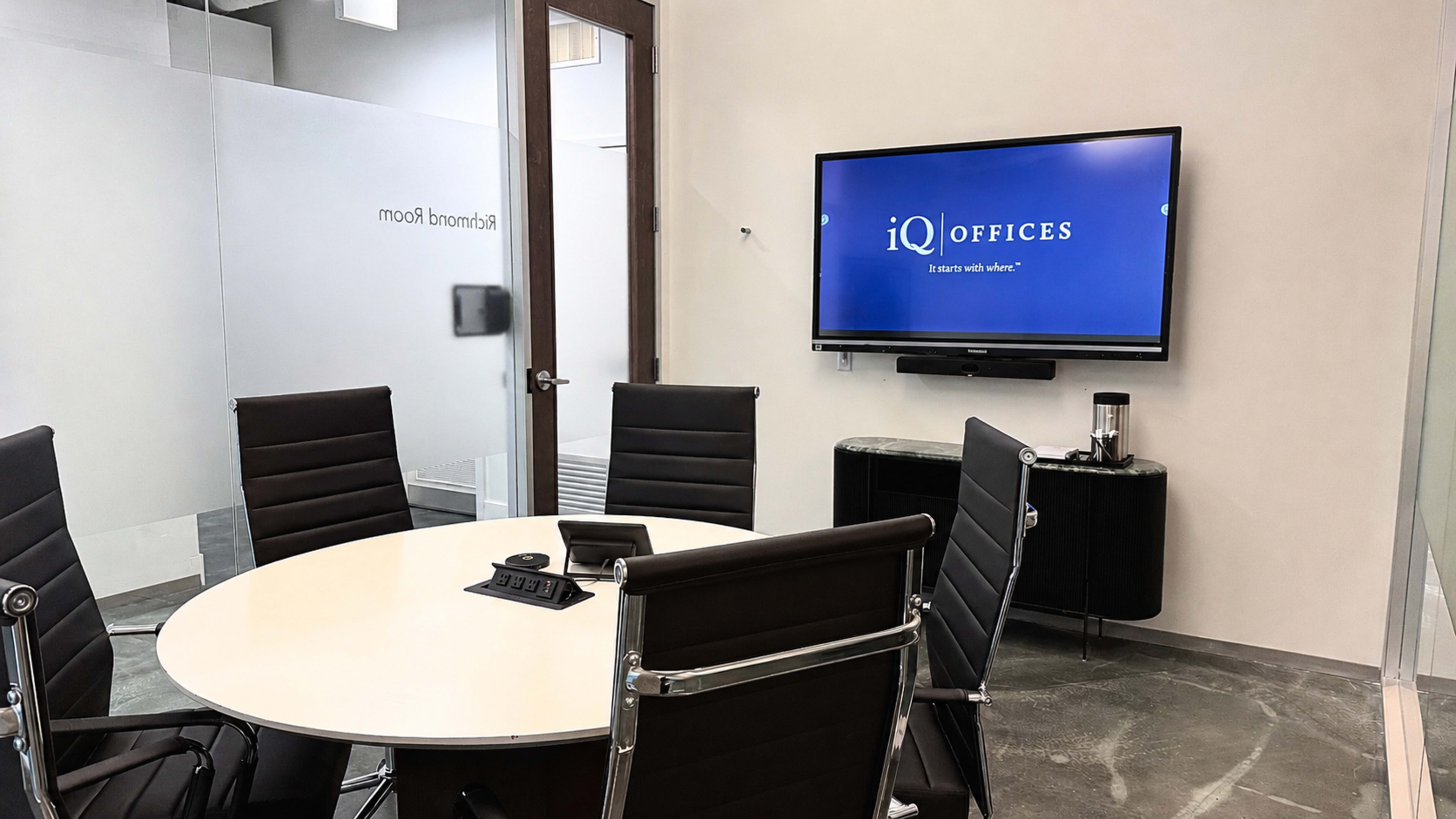 The image shows a modern conference room with a round table, black leather chairs, a wall-mounted screen displaying the iQ Offices logo, and a glass wall with a door labeled "Boardroom."