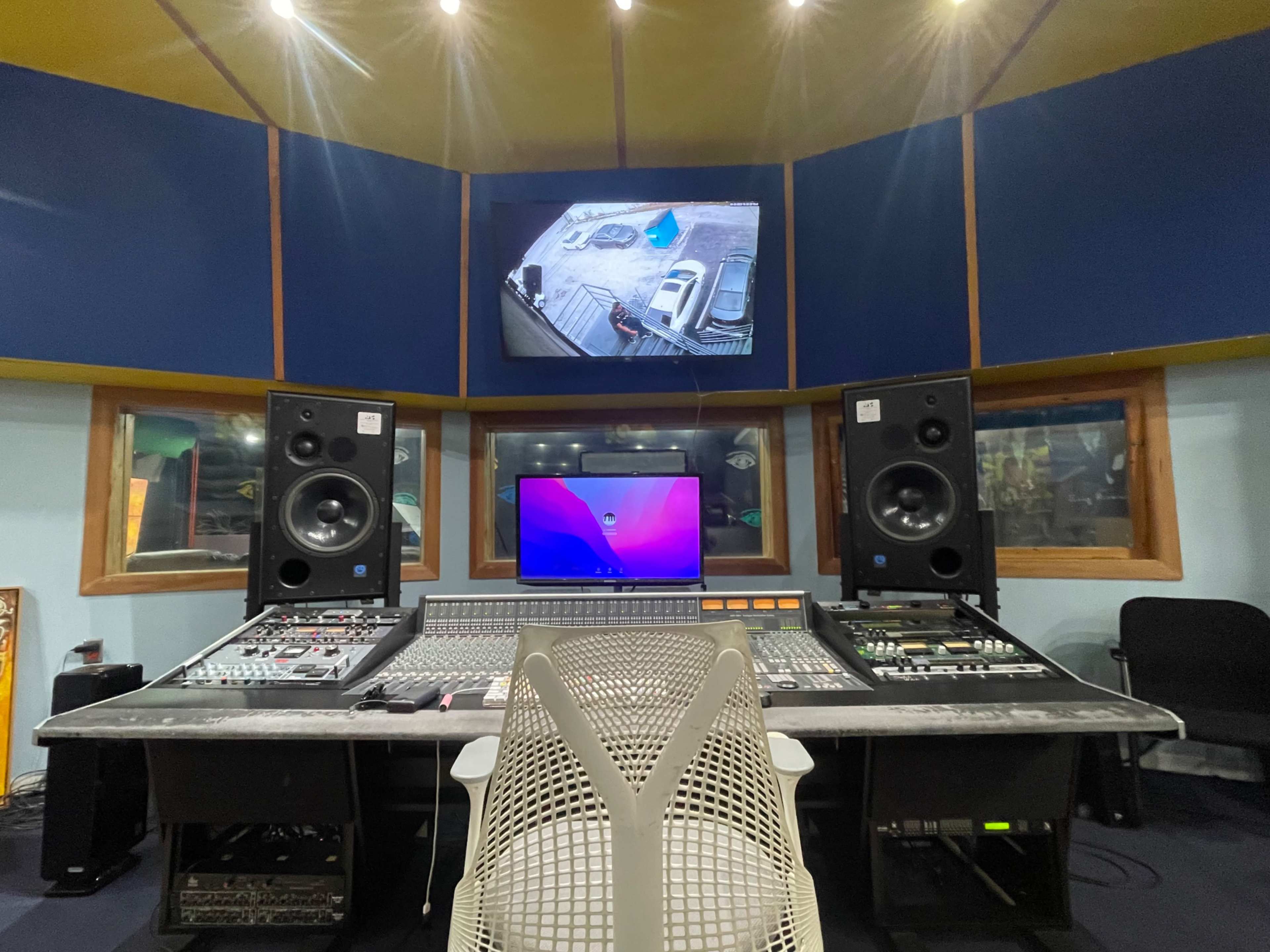 The image shows a music recording studio with a central mixing console, large speakers on either side, and a monitor displaying a video feed.