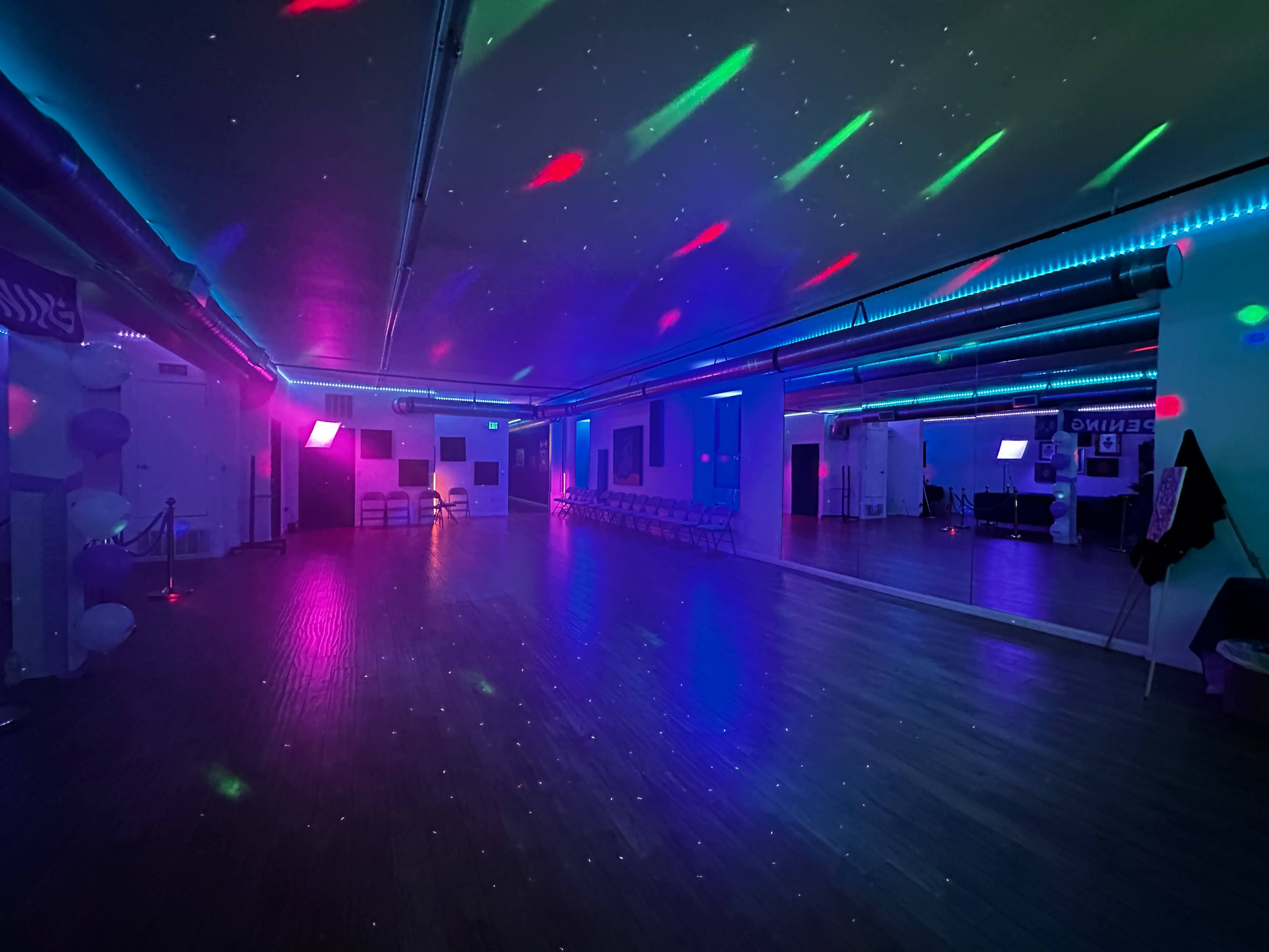 The image shows a dance studio with colorful lights reflecting off the walls and floor, creating a vibrant atmosphere.