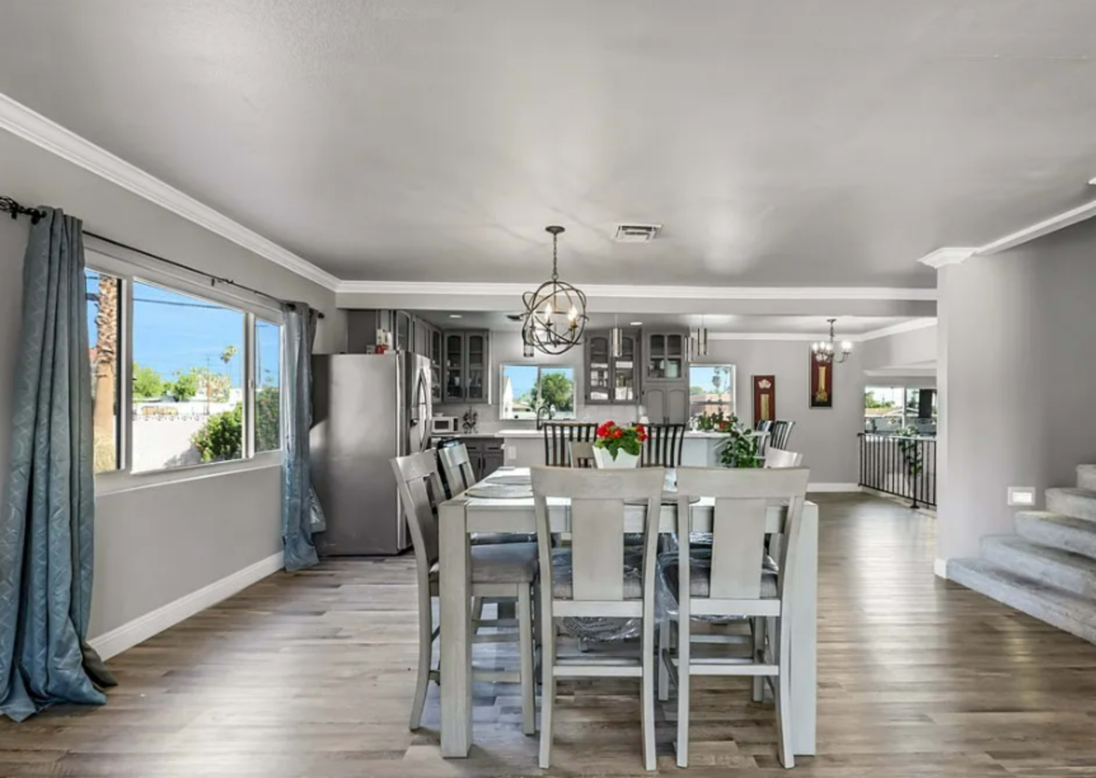 The image shows a modern dining area with a table and chairs, leading into a well-lit kitchen with gray cabinetry and a stainless steel refrigerator.