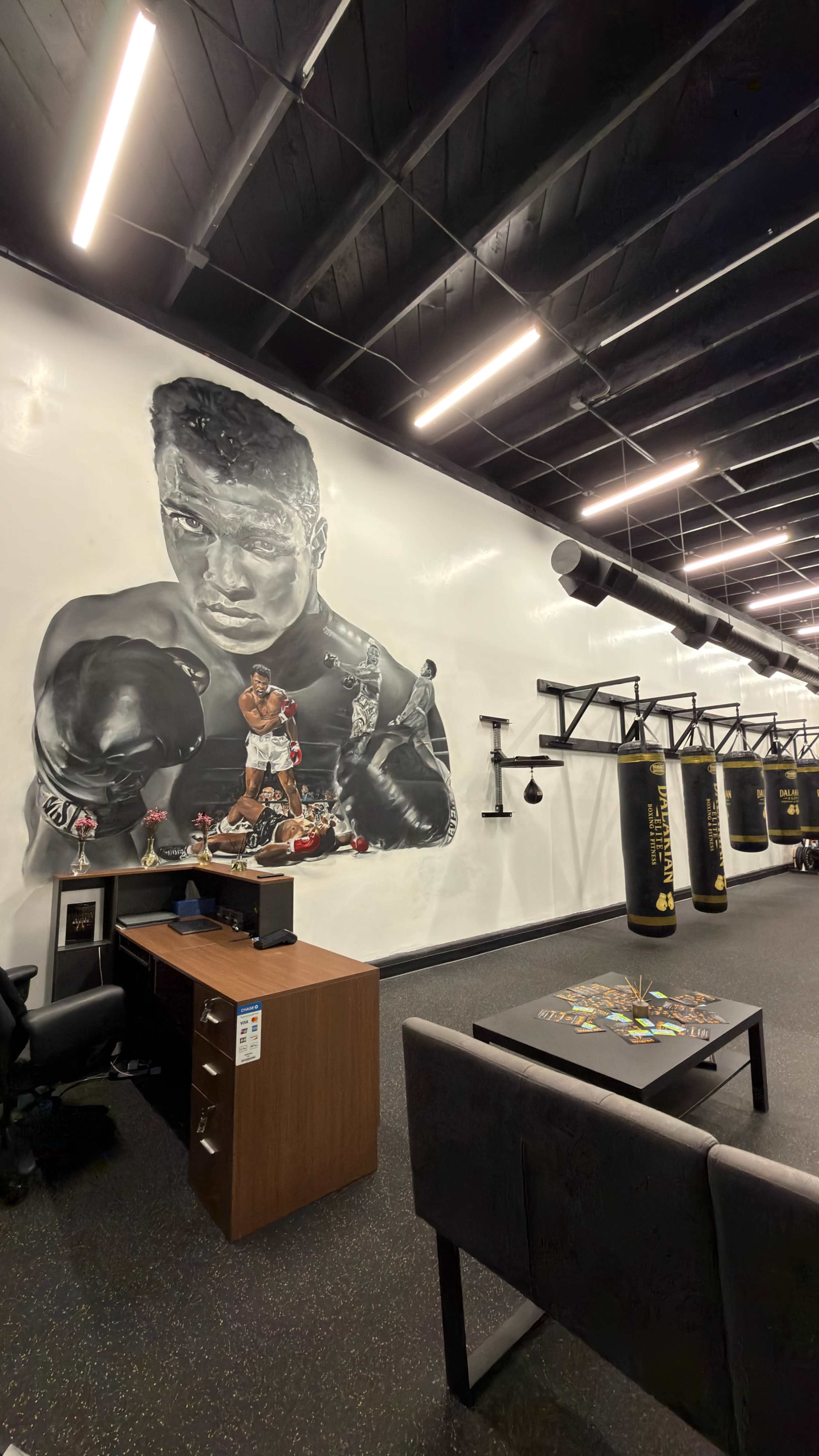 The image shows a boxing gym interior with a large mural of a boxer on the wall, punching bags hanging, and a desk with a computer in the foreground.