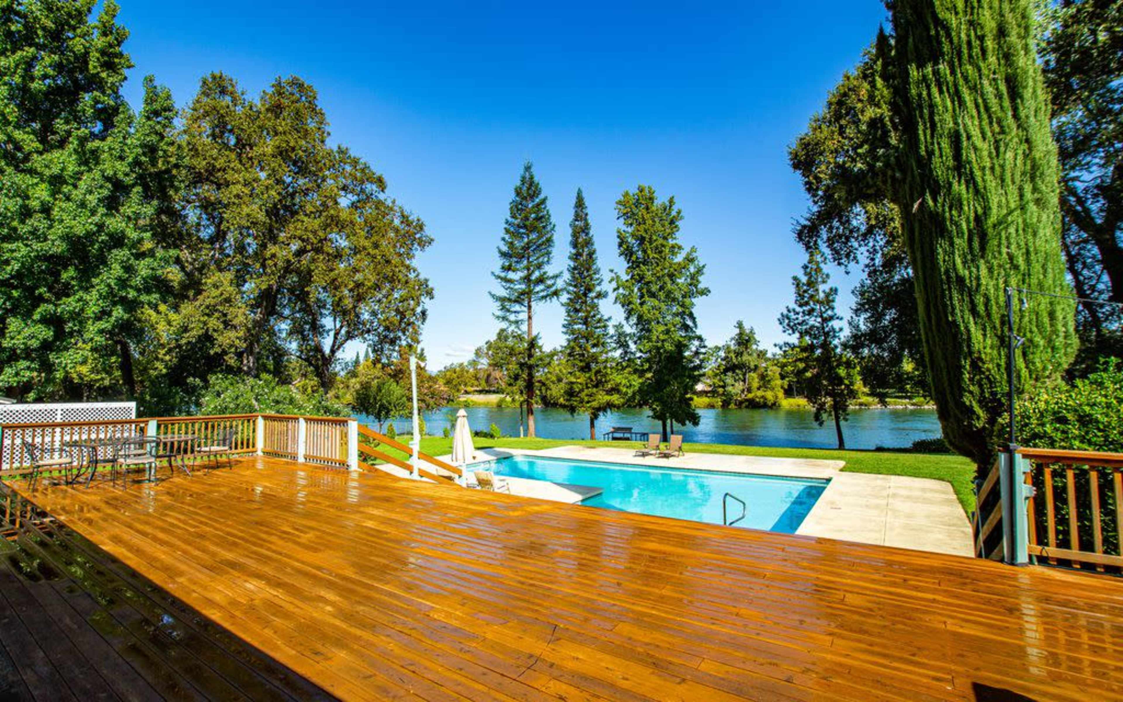 The image shows a wooden deck overlooking a swimming pool and a lake surrounded by tall trees under a clear blue sky.