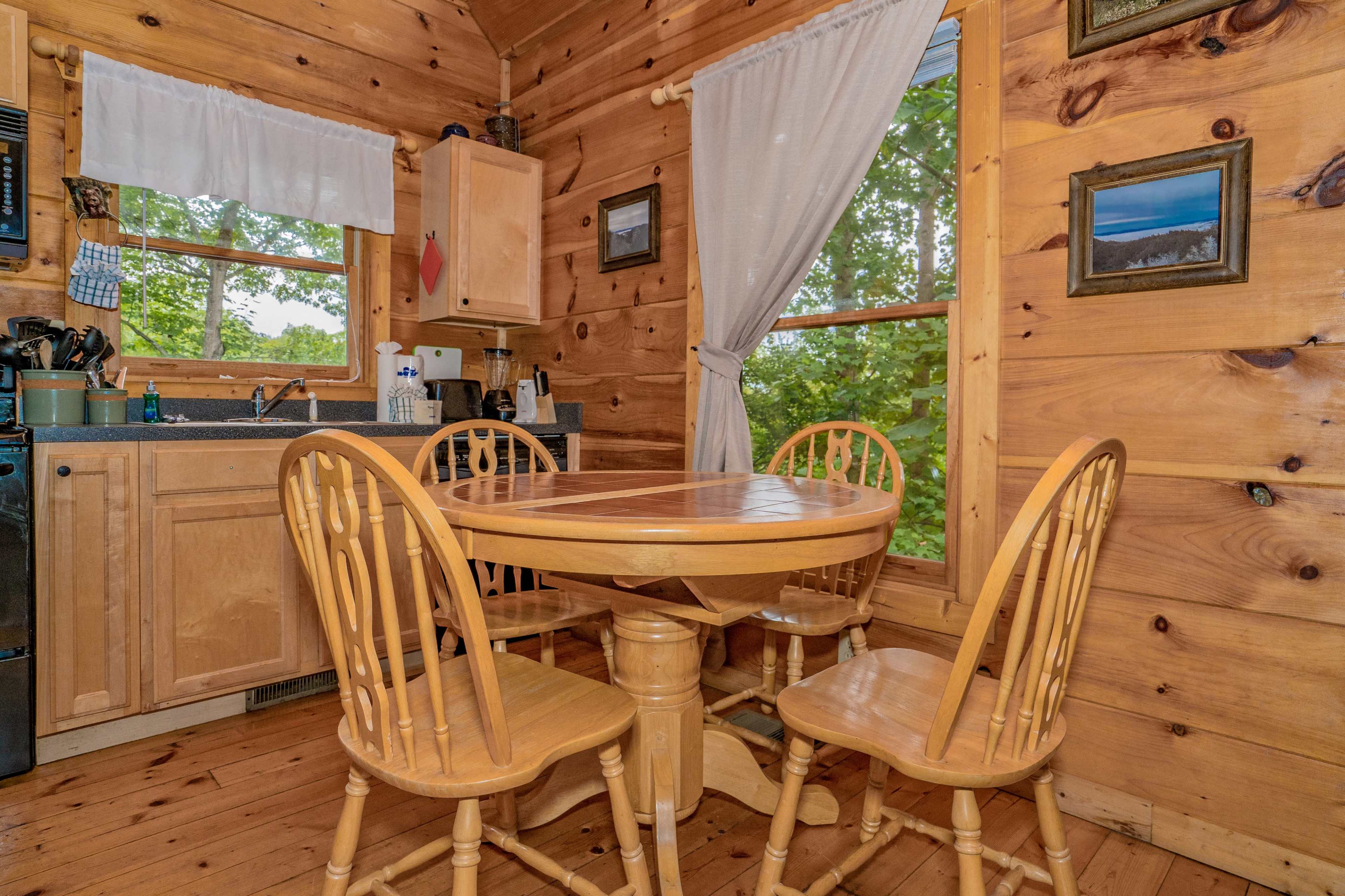 A wooden kitchen features a round table surrounded by four chairs, with a sink and cabinets visible in the background.