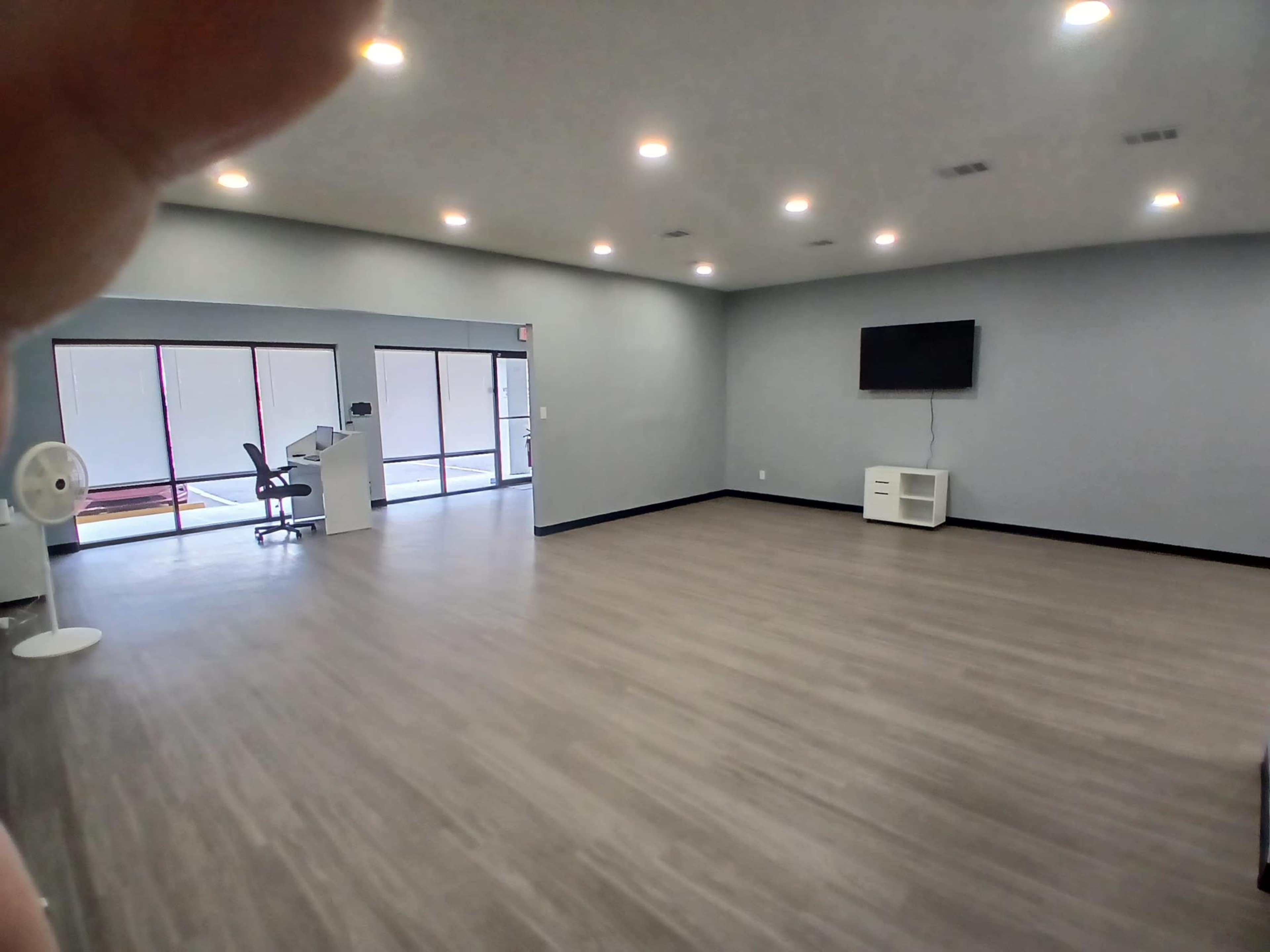 The image shows a spacious, empty room with wooden flooring, a large window, a wall-mounted TV, and minimal furniture including a fan and a desk chair.