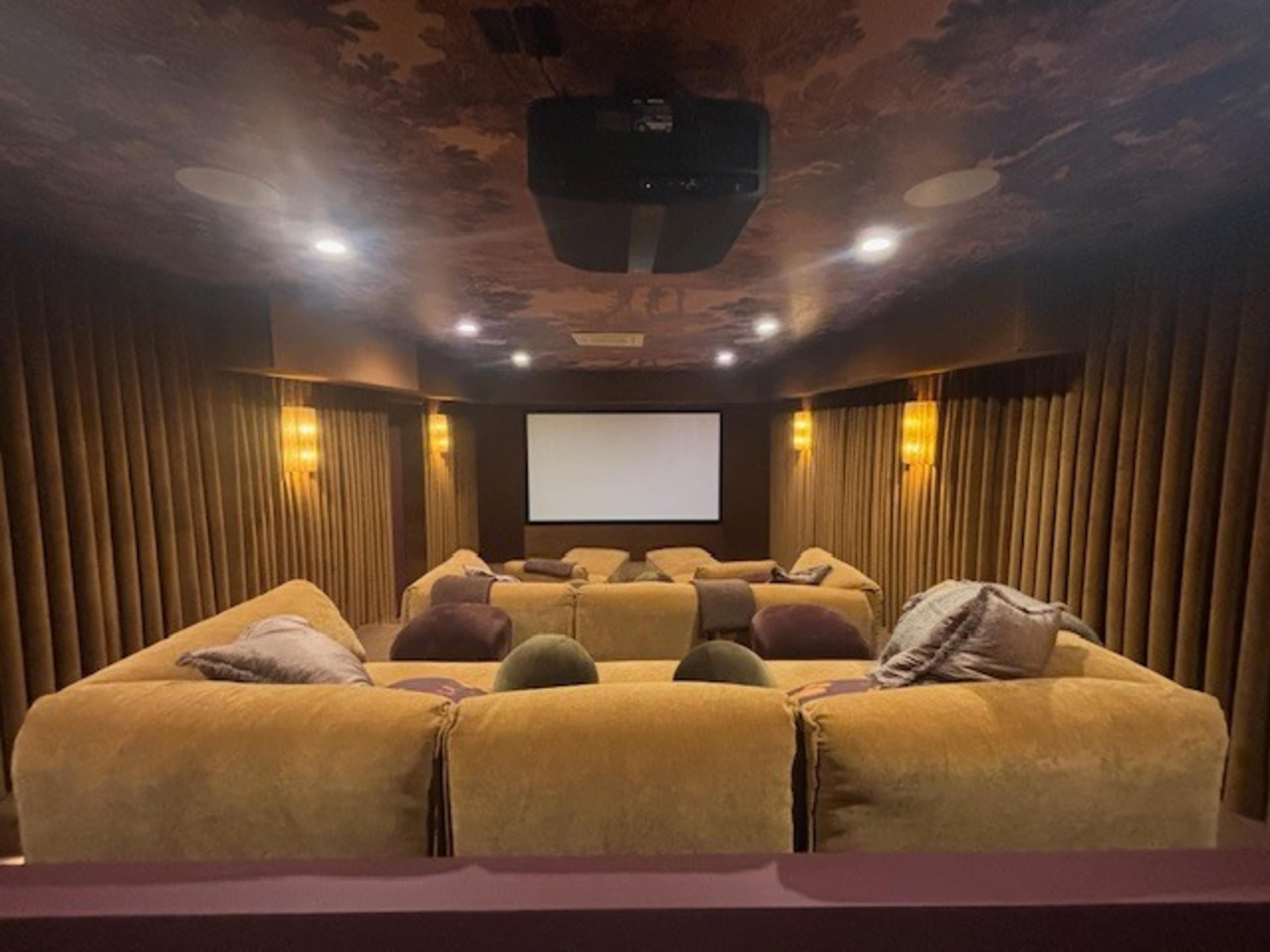 The image shows a dimly lit home theater with plush seating arranged in a semicircle facing a large screen, surrounded by curtains and wall lighting.