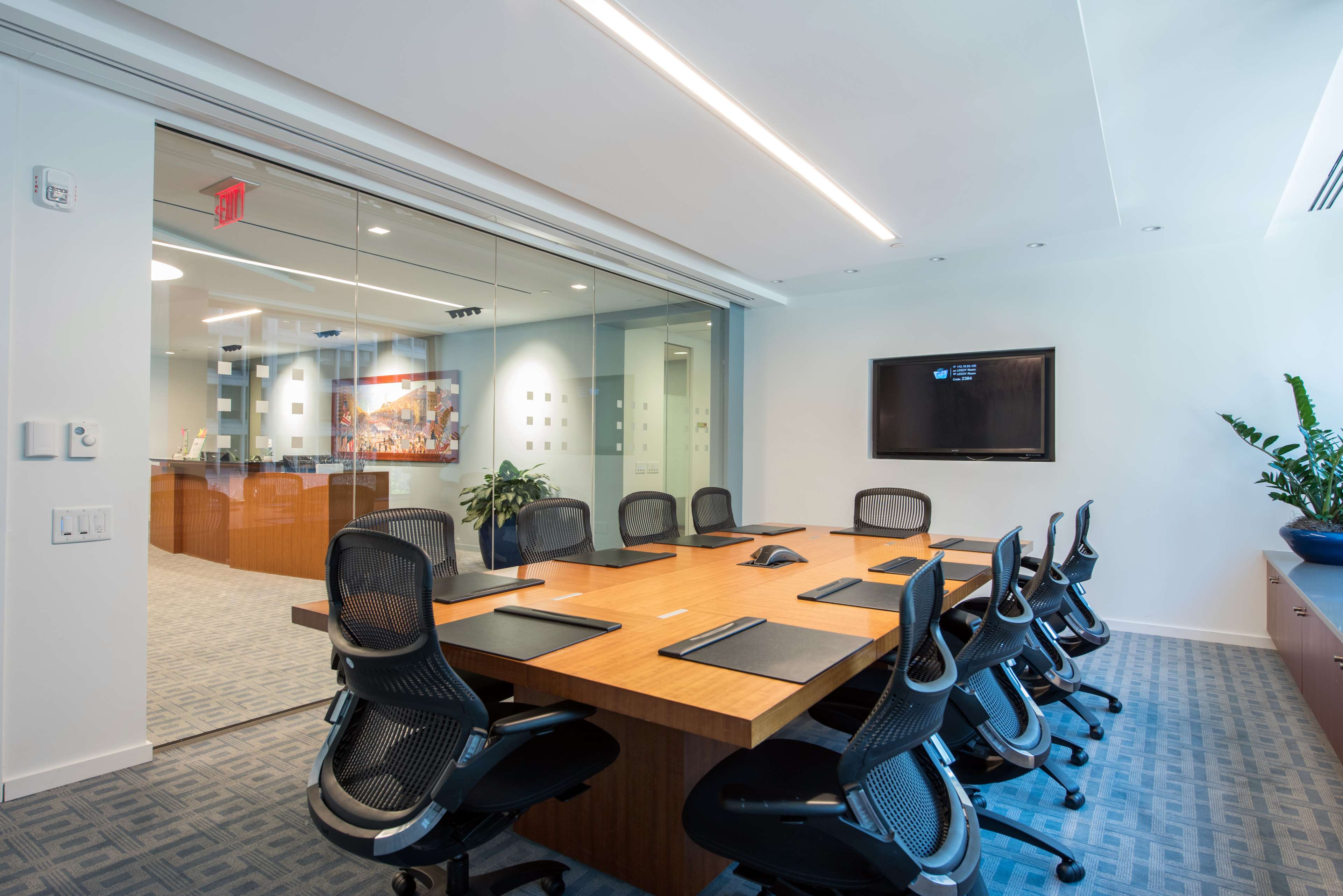A modern conference room features a large wooden table surrounded by ergonomic chairs, with a television on the wall and glass partitions.