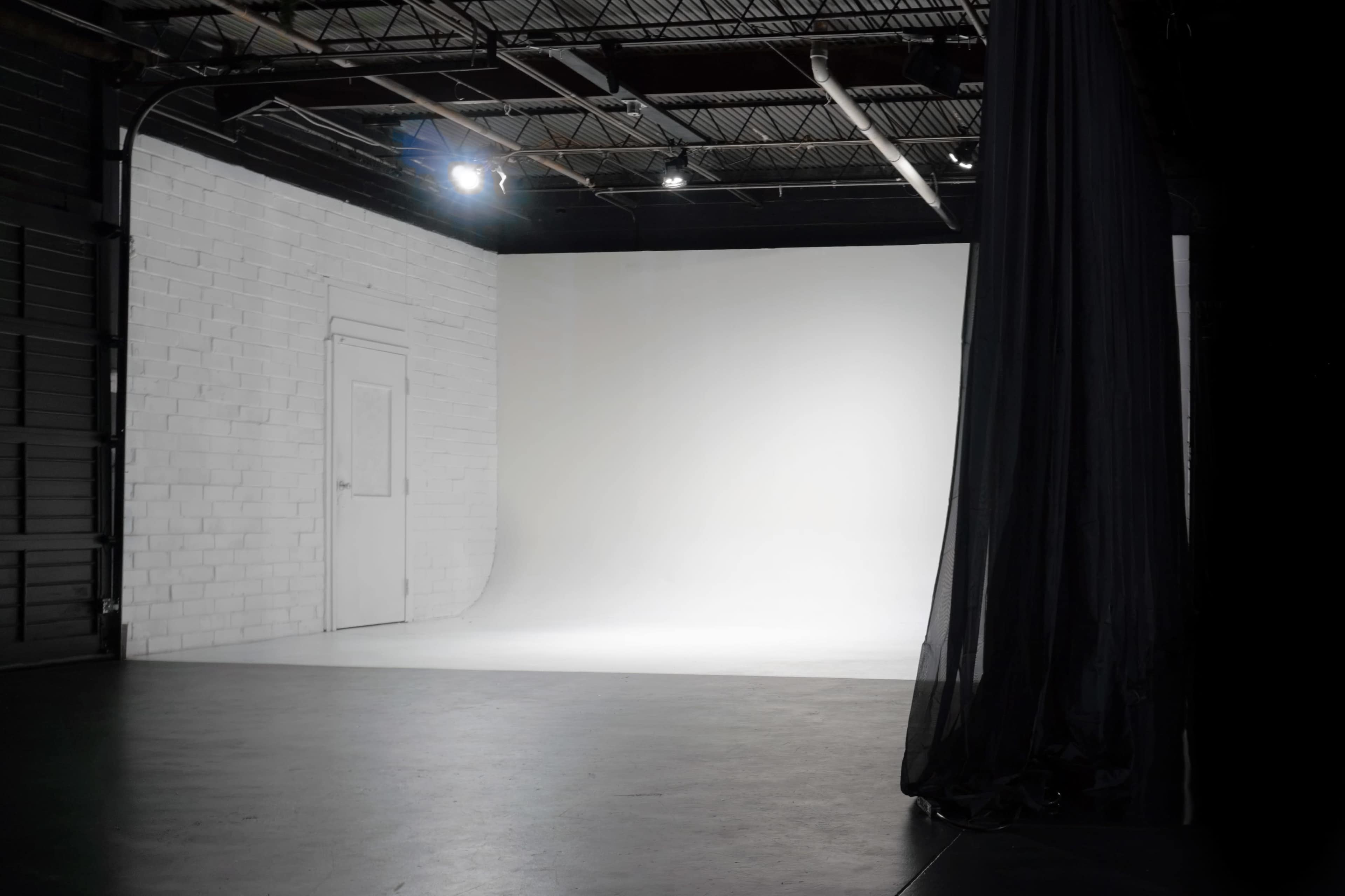 The image shows a minimalist studio space featuring a white brick wall, a closed door, and a large, empty white backdrop.