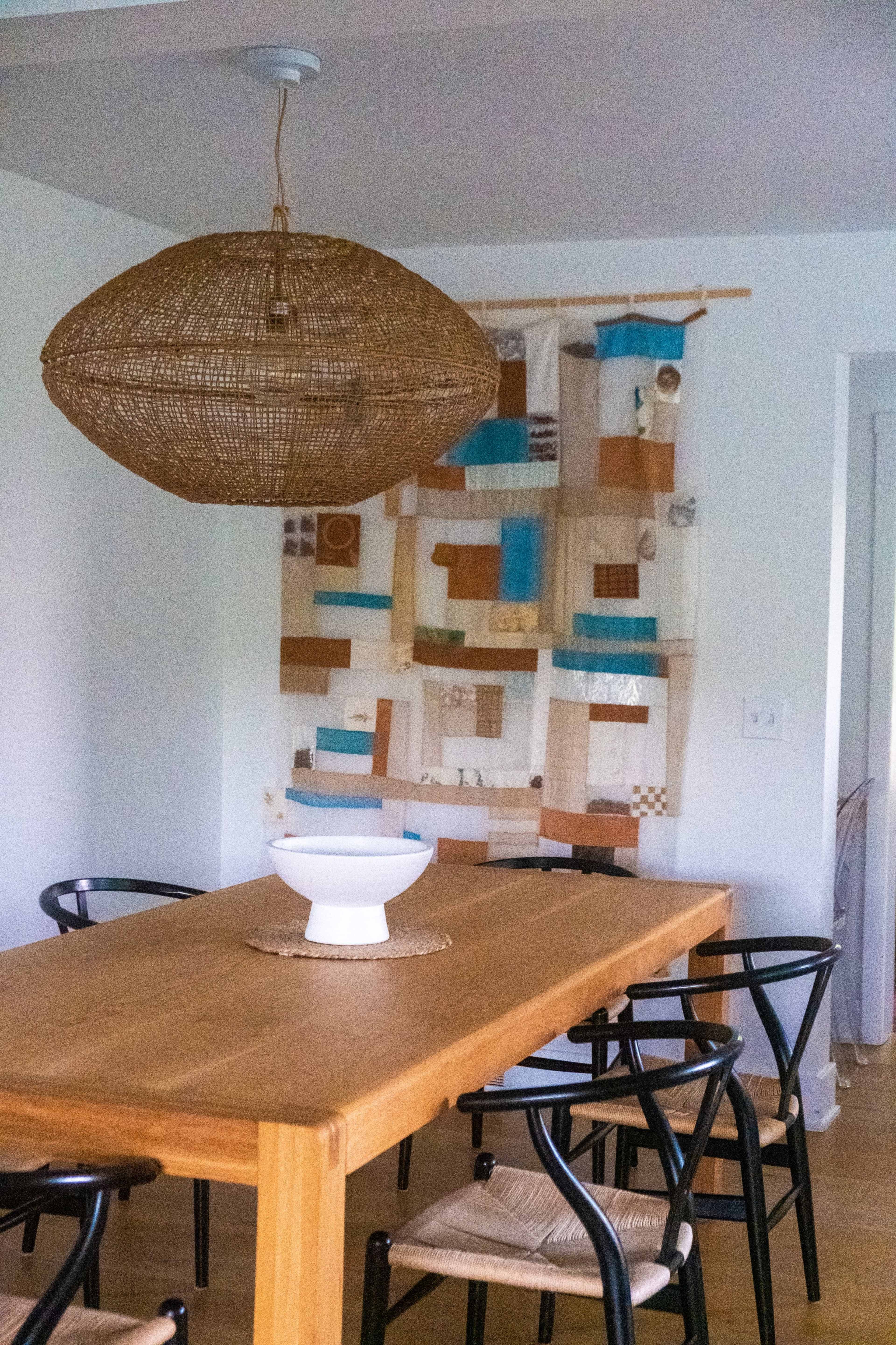 A wooden dining table is surrounded by black chairs, with a woven pendant light above and a textile wall hanging behind it.