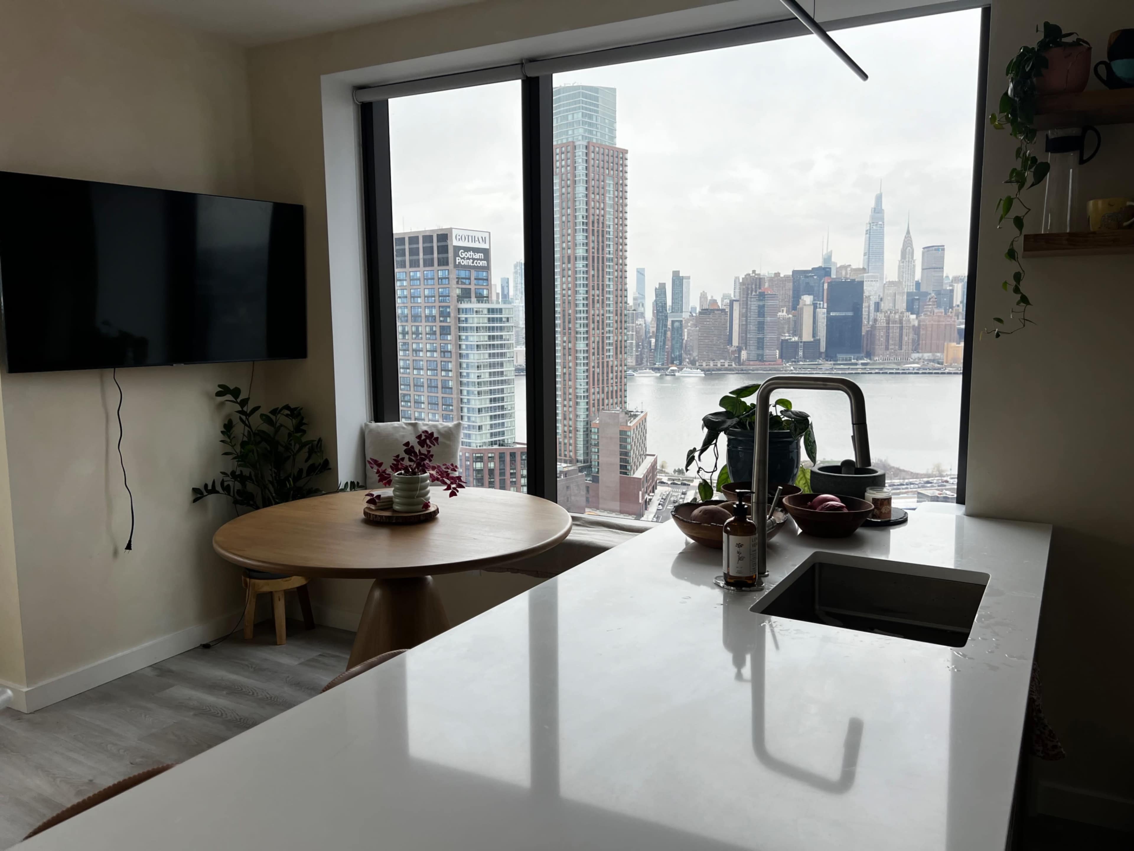 A bright kitchen features a modern countertop and sink, with a round dining table nearby, overlooking a city skyline through a large window.