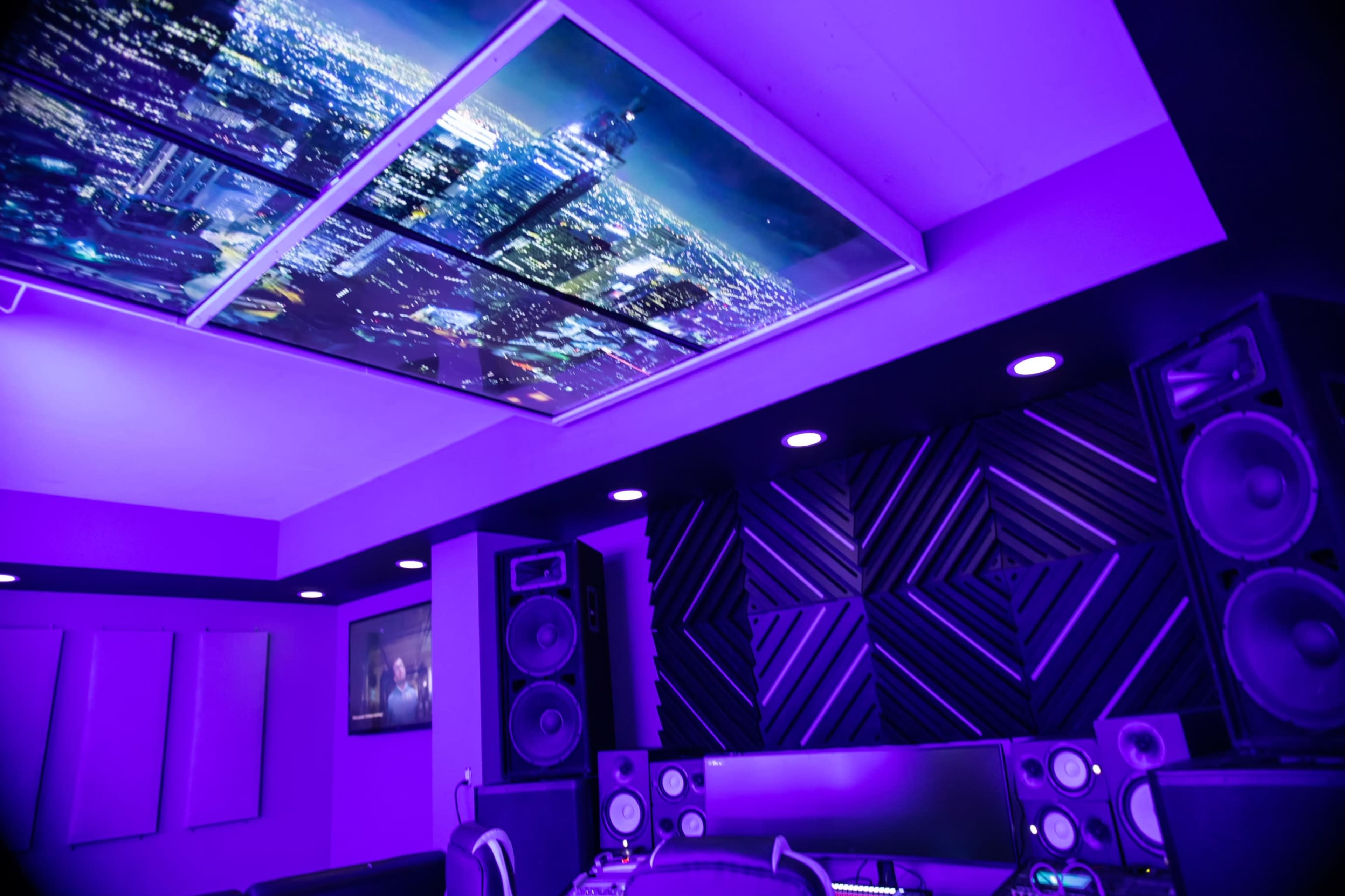A music studio features purple lighting, large speakers, and a ceiling display of a city skyline at night.