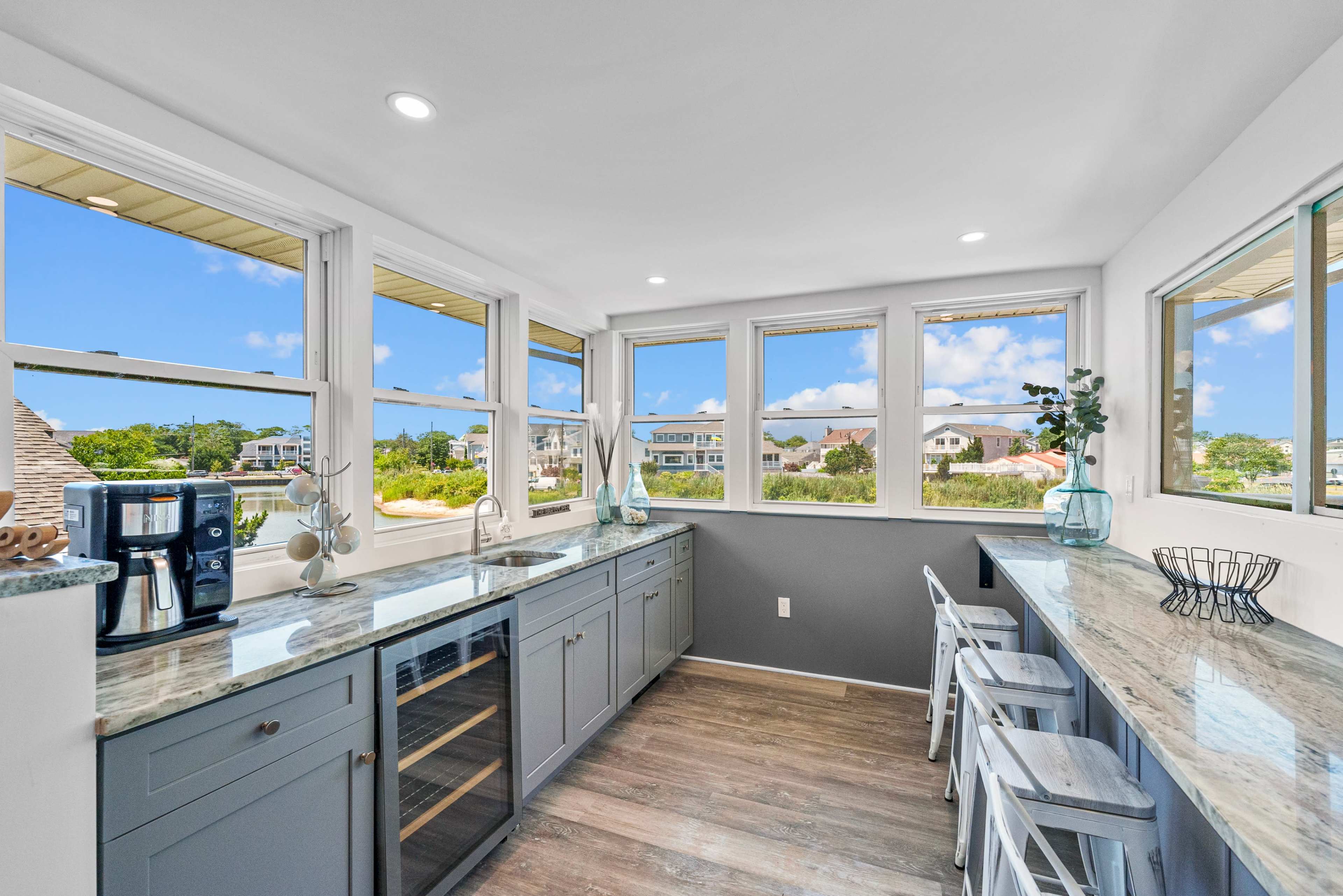 The image shows a bright kitchen with large windows, light-colored cabinetry, and a countertop with bar seating, overlooking a scenic view.