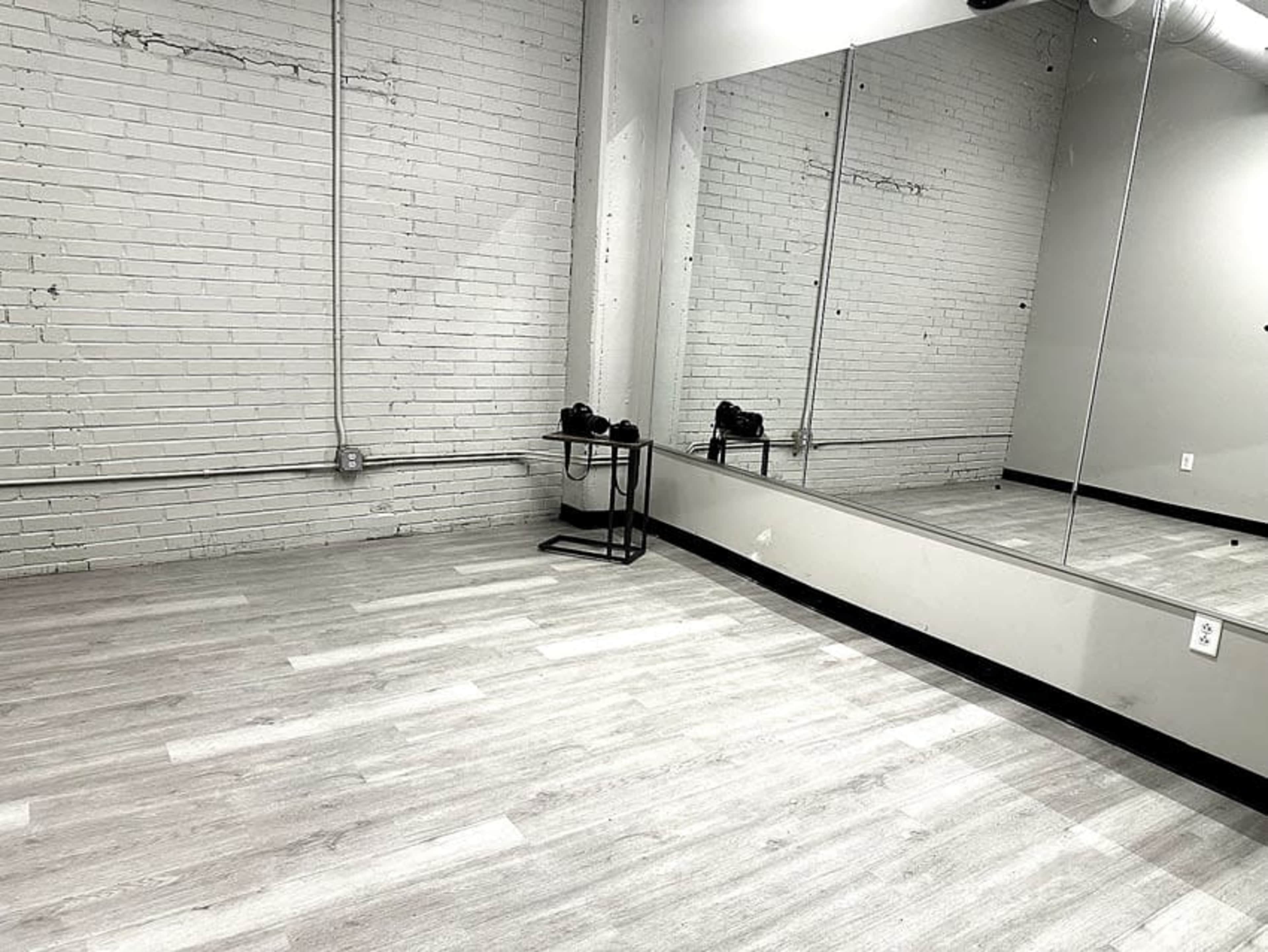 The image shows a spacious, empty room with white brick walls, a large mirror, and light-colored flooring, featuring a small table with two objects on it.