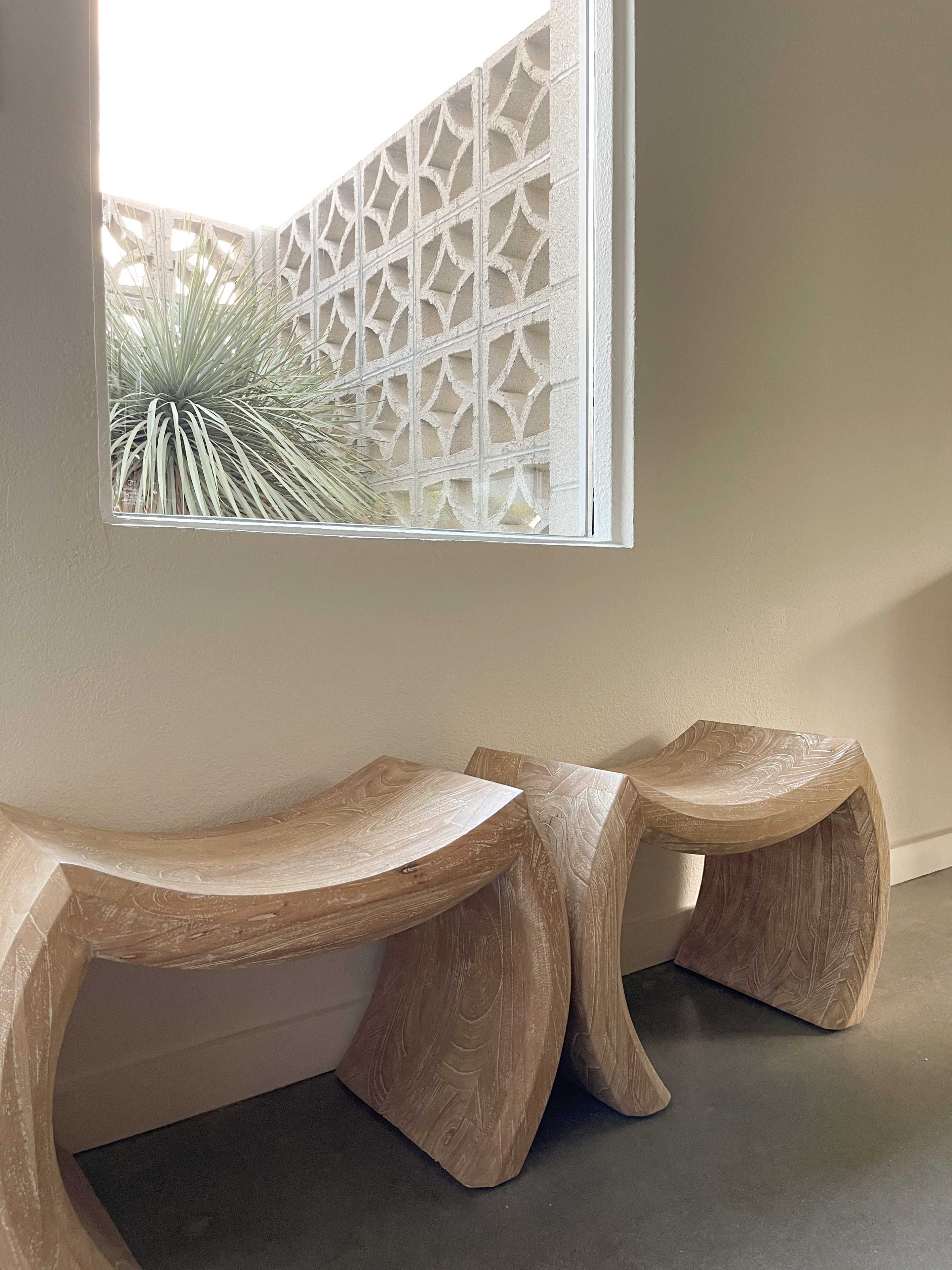 Two curved wooden stools are placed next to a window that frames a geometric wall and a spiky plant outside.