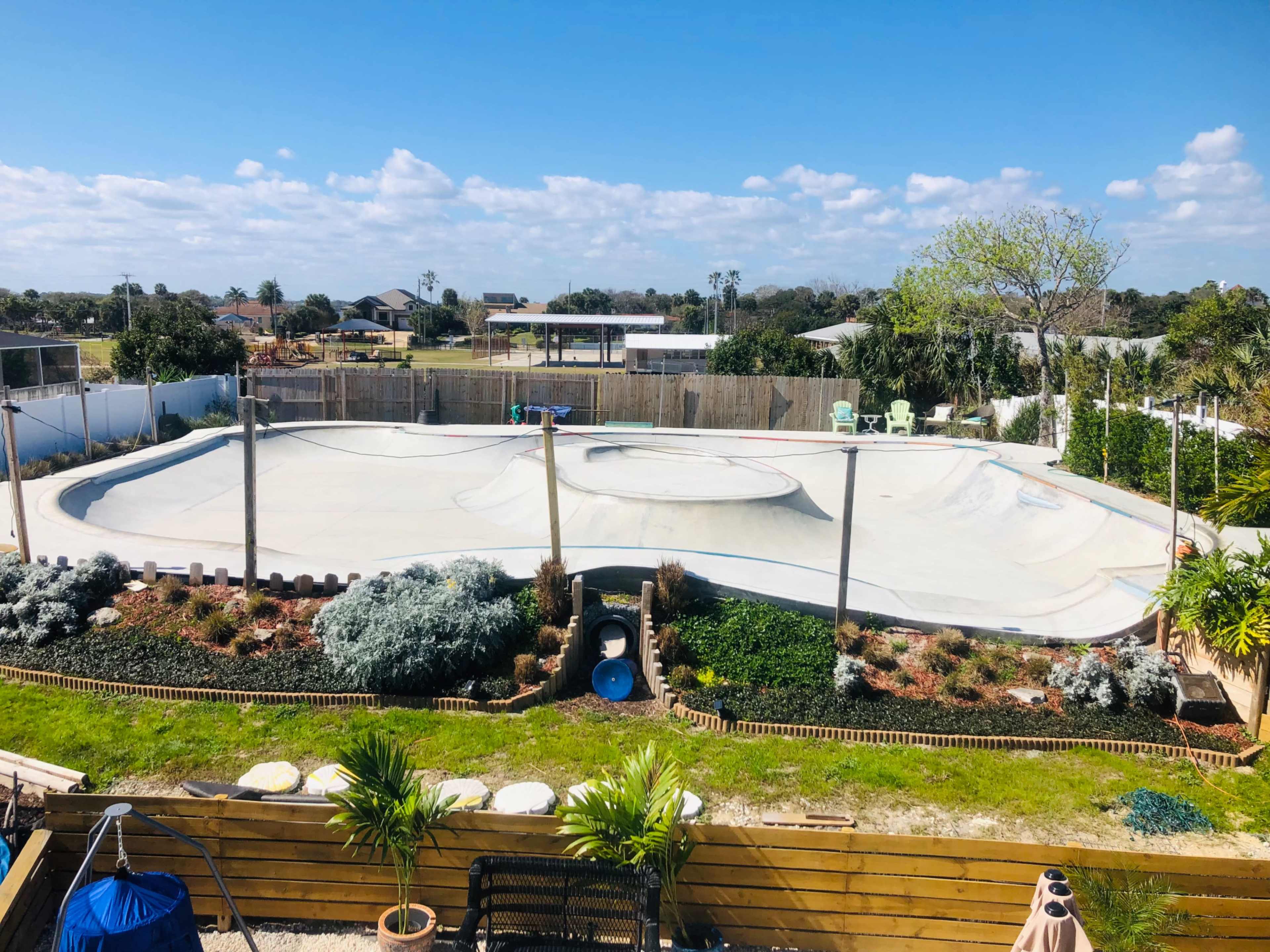 The image shows a large, empty skatepark with a concrete surface surrounded by landscaped gardens and a few trees under a clear blue sky.