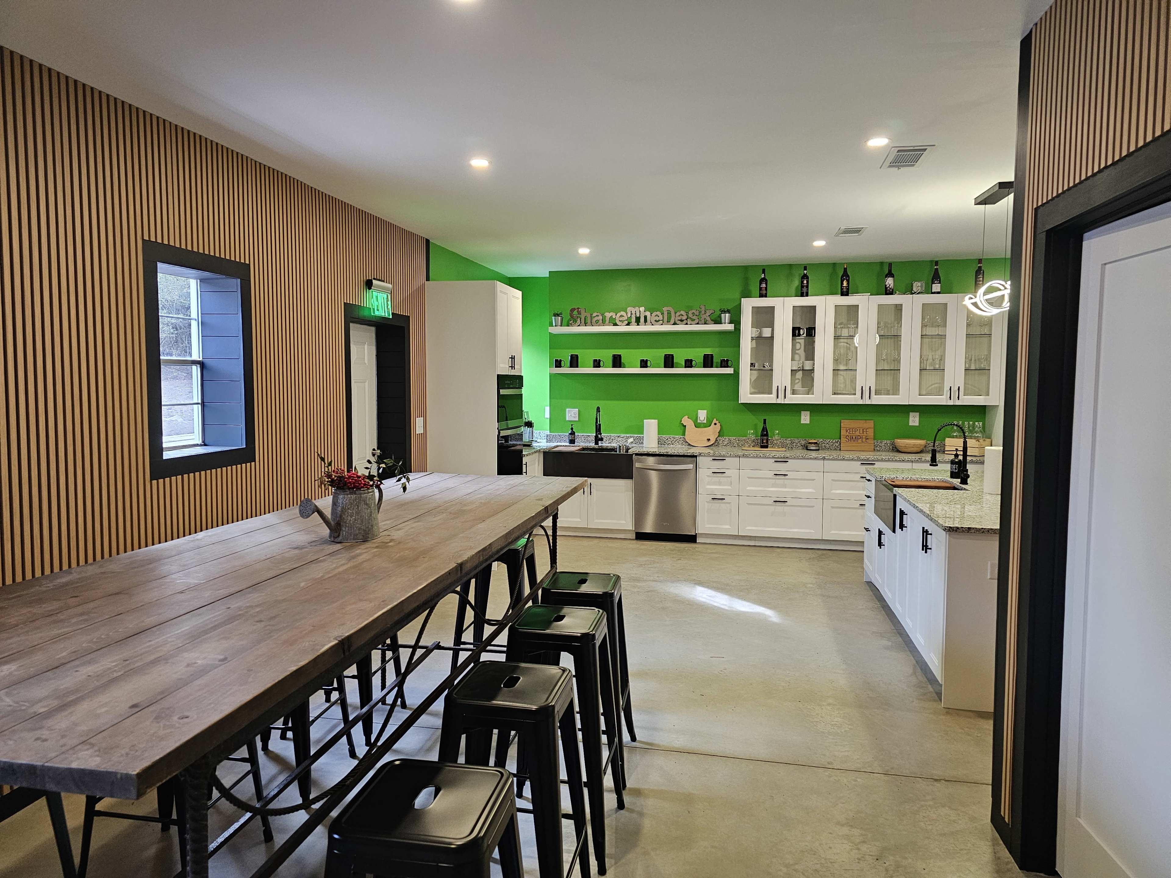 The image shows a modern kitchen with white cabinets, a large wooden table with black stools, and a vibrant green accent wall.