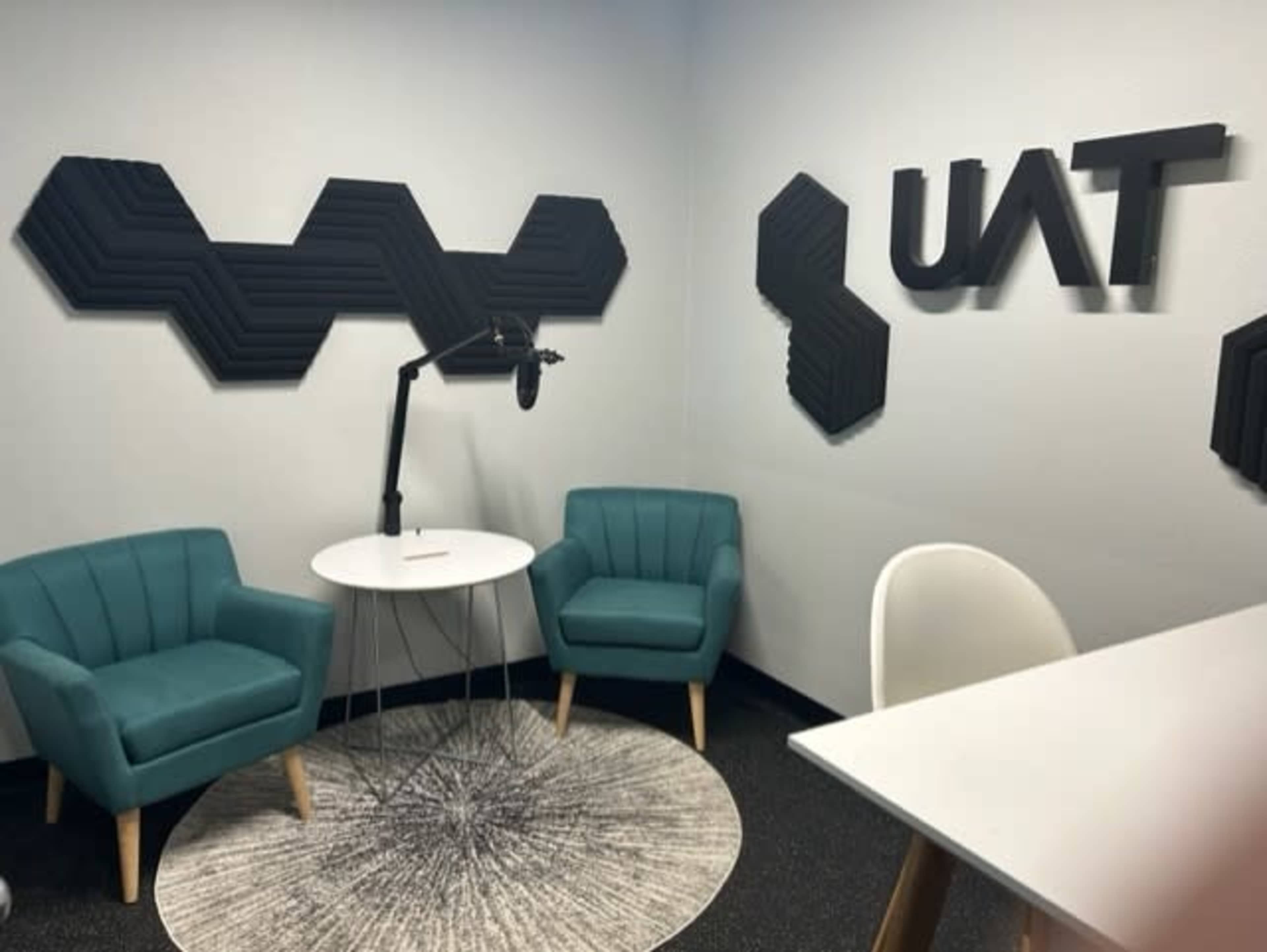 A modern room with two teal chairs, a round table, and a microphone stand, featuring acoustic wall panels and the letters "UAT" on the wall.