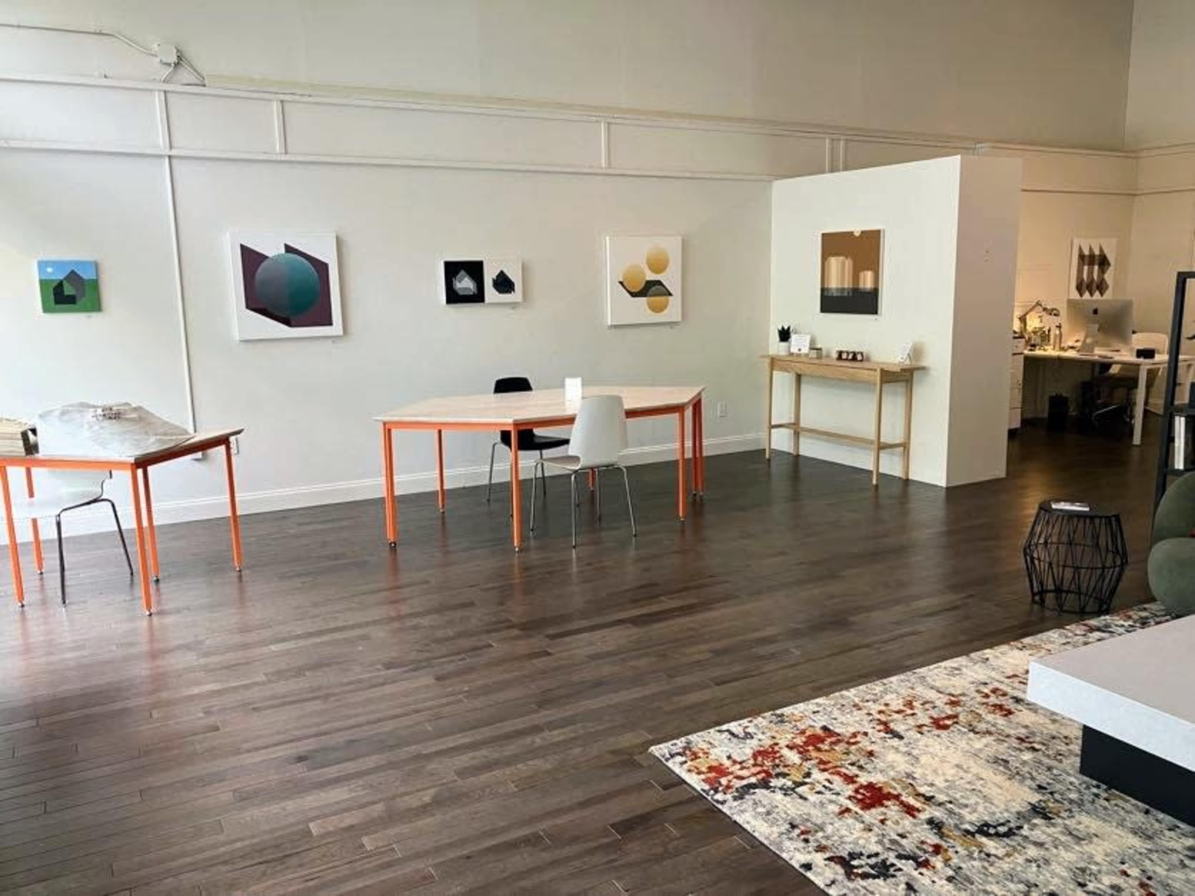 An art gallery space with wooden flooring, featuring abstract artworks on the walls and a couple of tables with chairs arranged in the foreground.