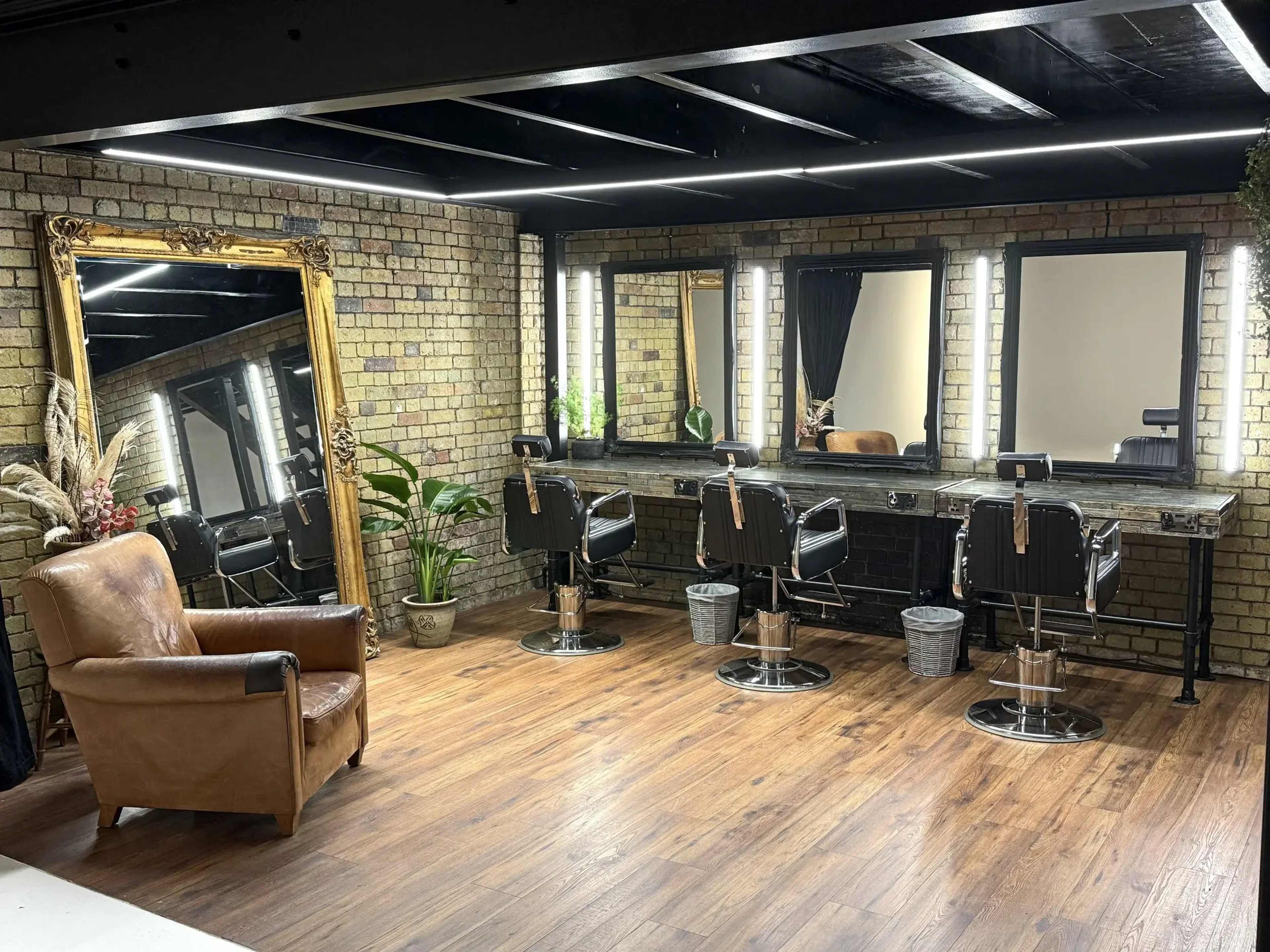 The image shows a modern hair salon interior featuring four styling stations with mirrors, a vintage chair, and decorative plants against exposed brick walls.