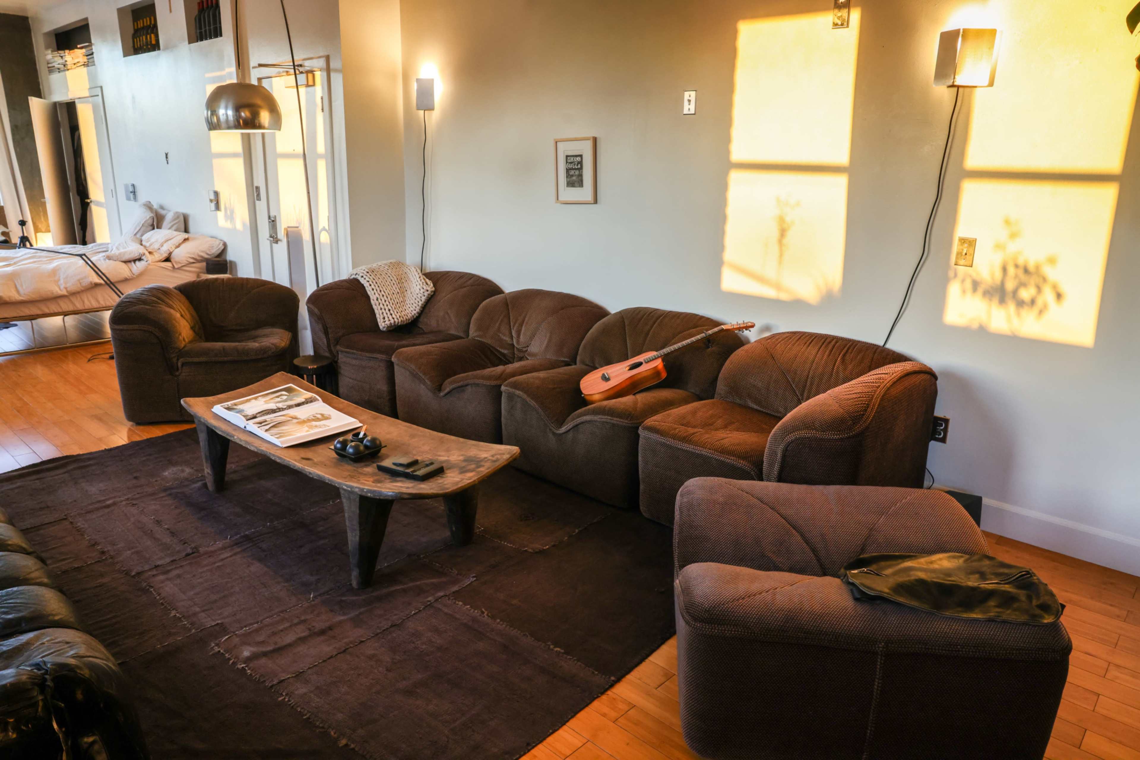 The image shows a living room with a brown sofa set, a wooden coffee table, and sunlight casting shadows on the wall.