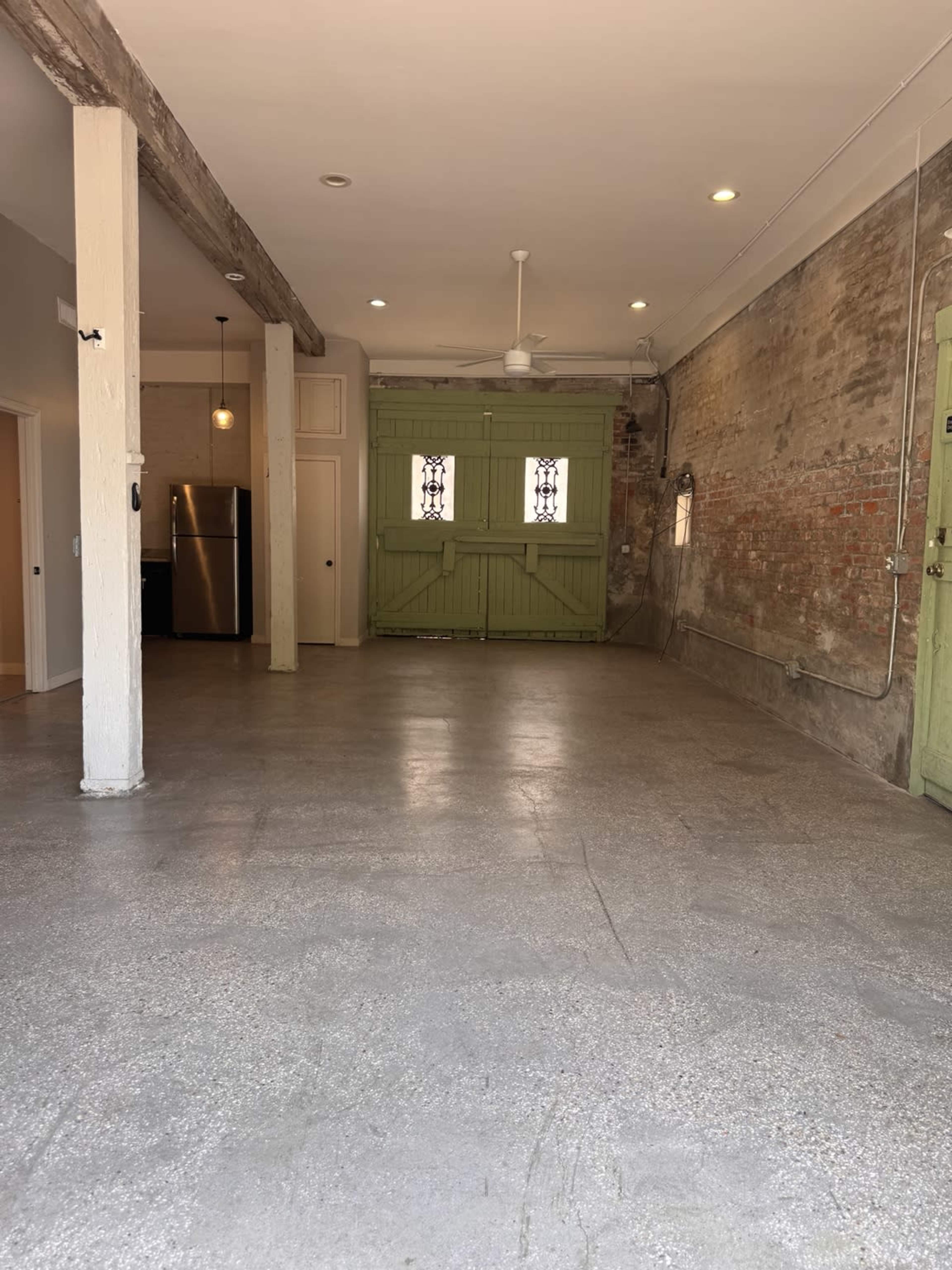 The image shows an empty interior space with exposed brick walls, a concrete floor, and a green double door at the far end.
