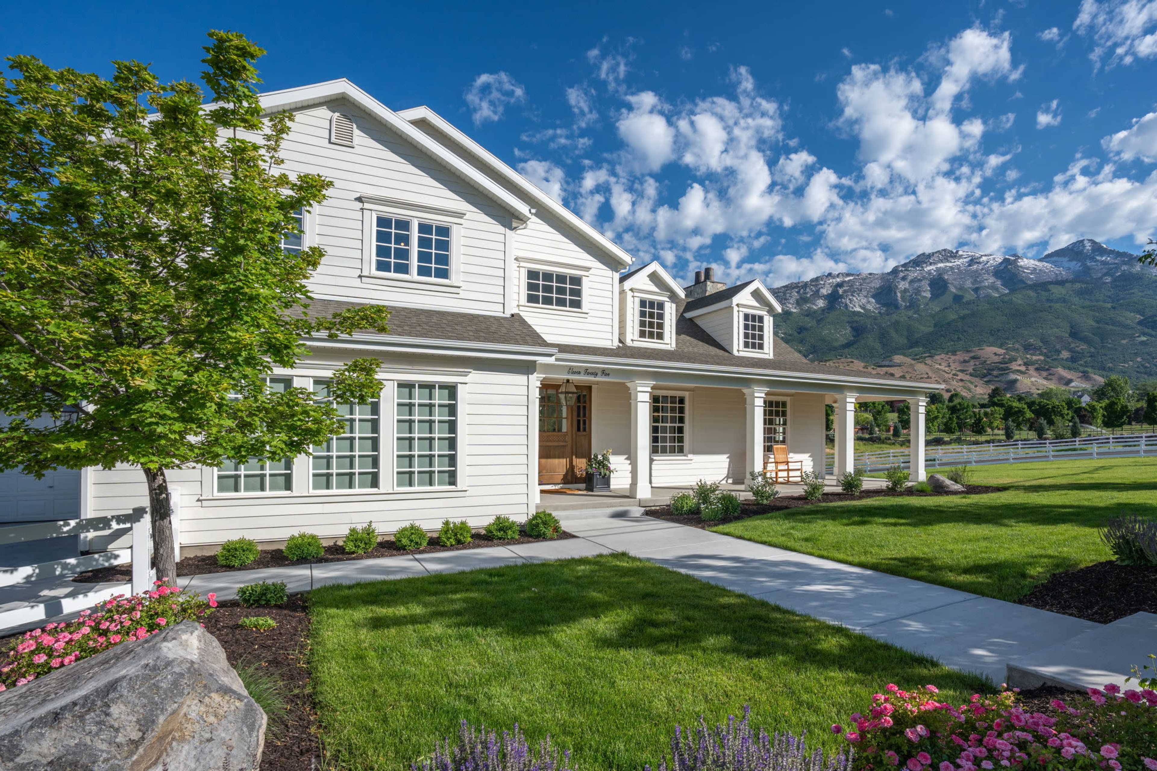A well-maintained two-story white house with a large front porch is set against a backdrop of mountains and a blue sky.