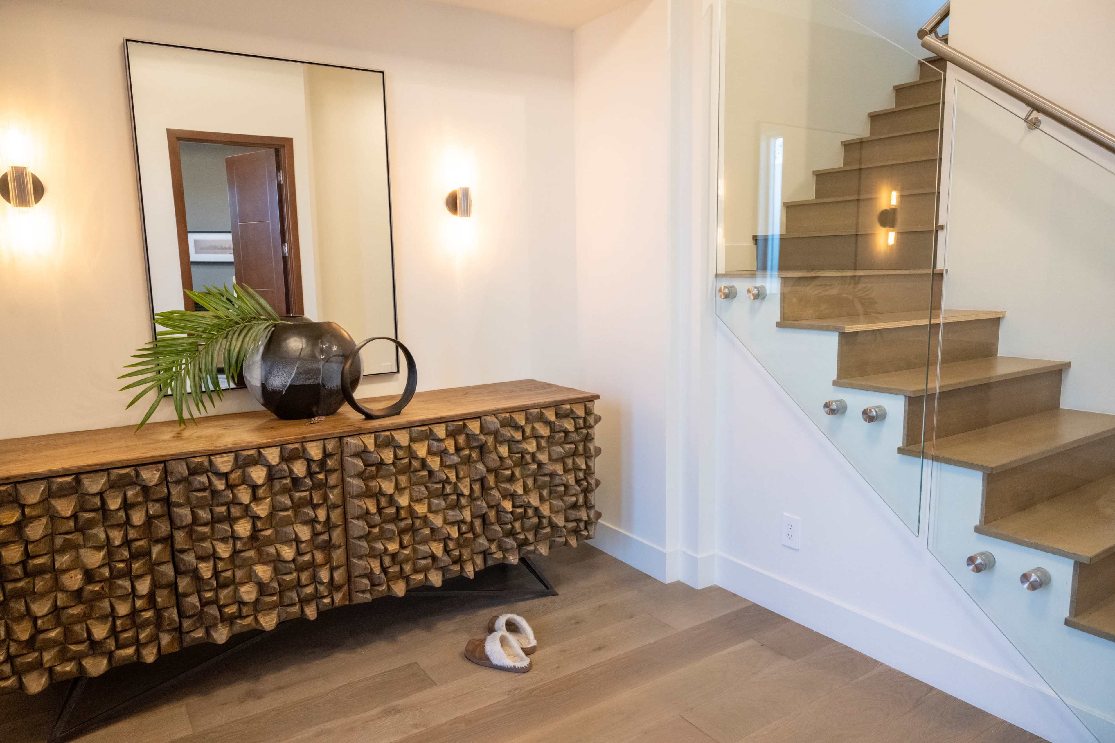 An entryway with a wooden sideboard featuring a textured design, a large black vase with greenery, a mirror on the wall, and a staircase with glass railing leading to the upper level.