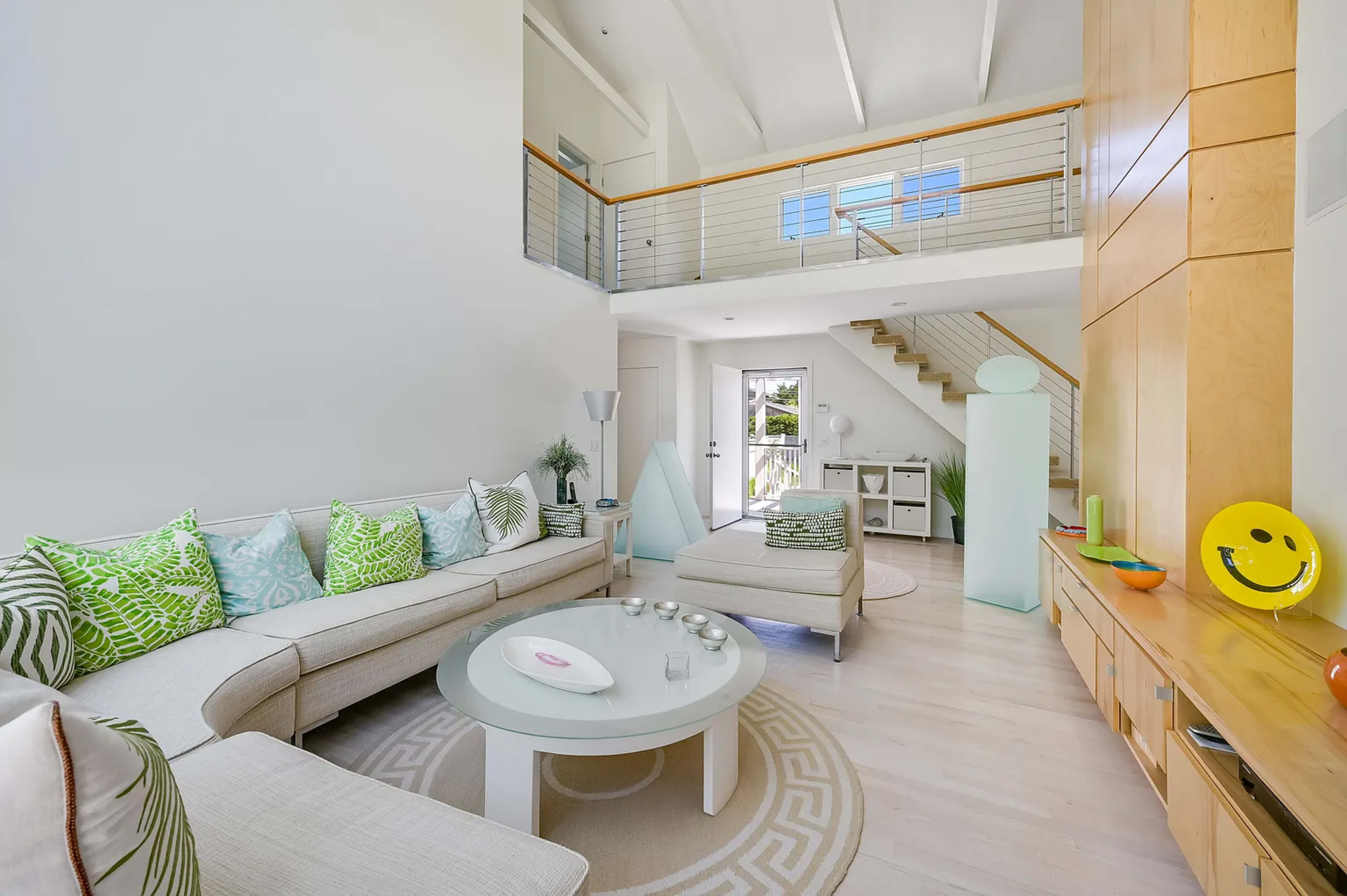 A modern living room with a white couch, patterned cushions, a glass coffee table, and a staircase leading to an upper level.