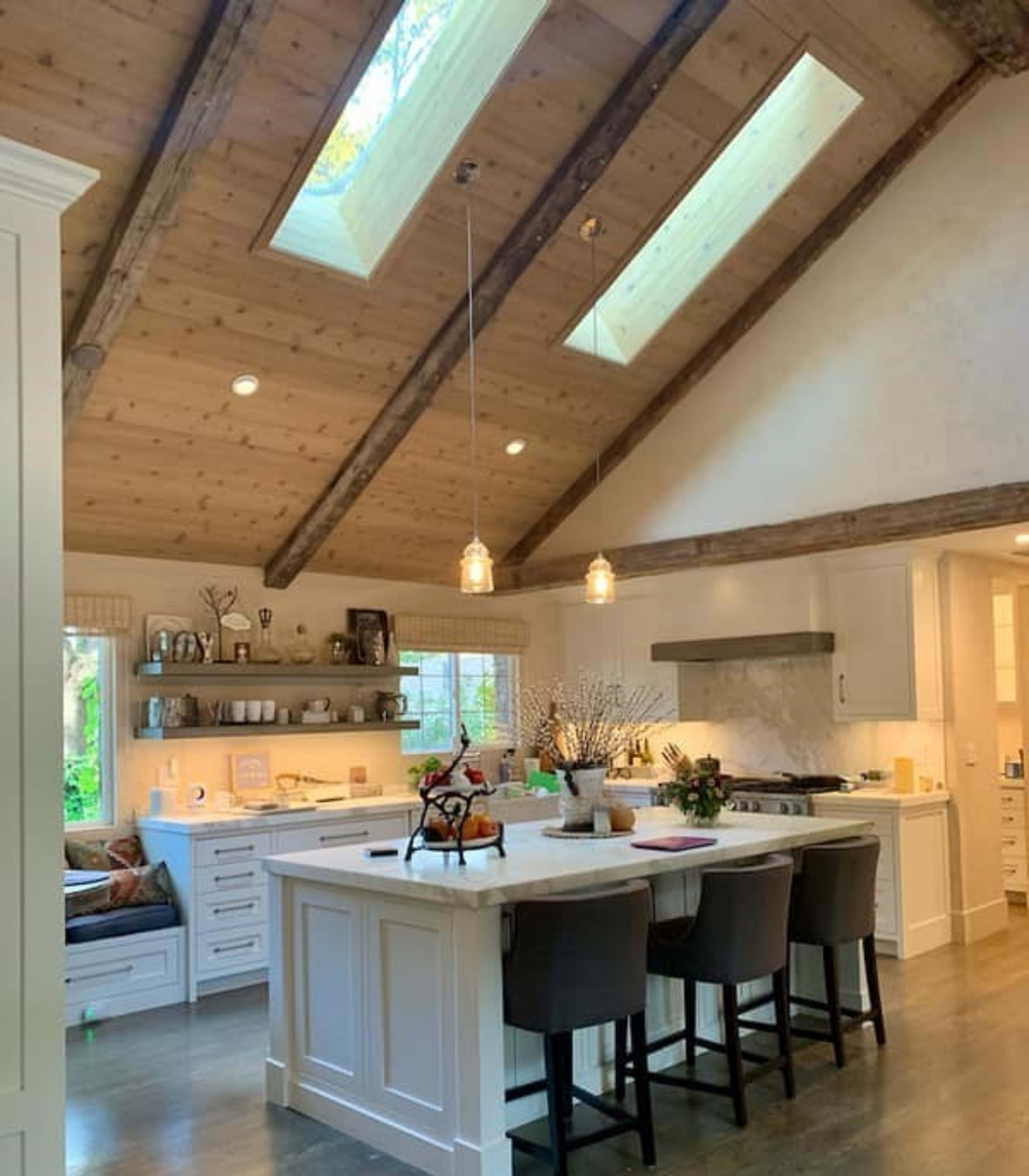 A modern kitchen with a central island, wooden beams, skylights, and a cozy seating area by a window.